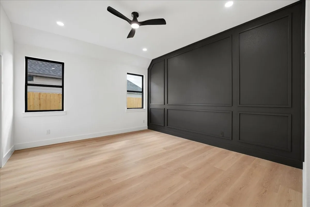 Wood-finish flooring, accent wall with panel molding, two windows with black trim, recessed lighting, and a ceiling fan