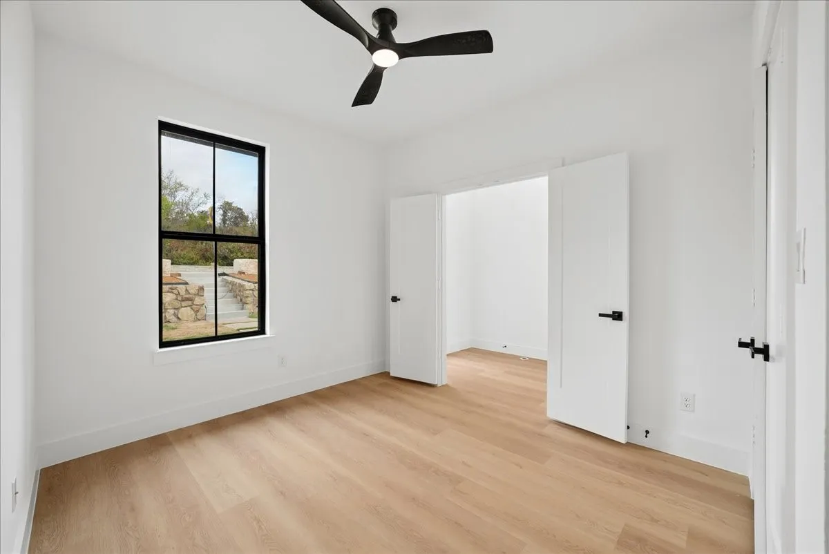 Light wood-finish flooring, white walls, black-framed window, black ceiling fan, and white doors with black hardware