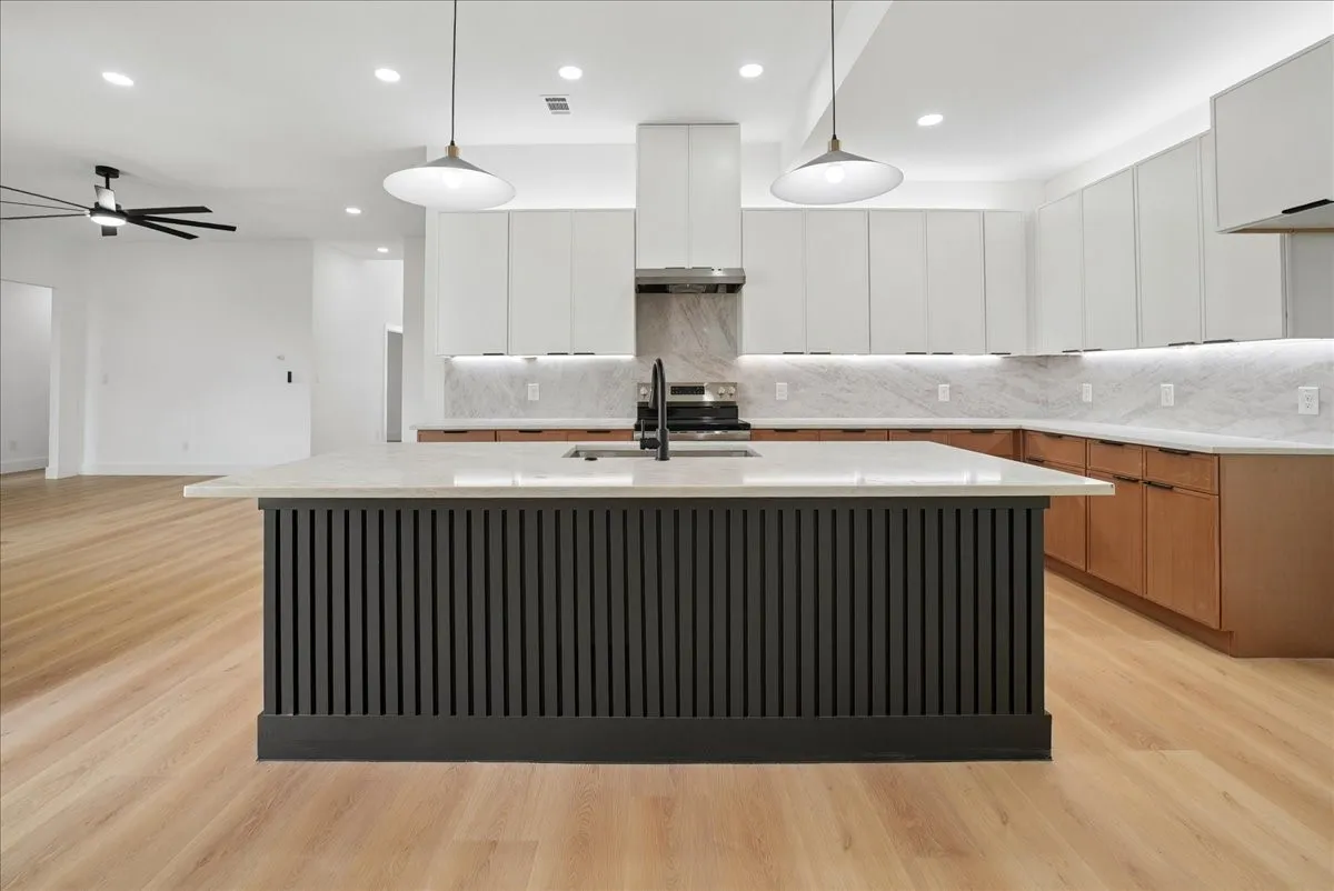 Contemporary kitchen featuring a striking black vertical-slat island with waterfall countertop edge, white shaker cabinetry, subway tile backsplash, stainless steel appliances including a professional-style range hood, warm wood-finish flooring, and pendant lighting suspended above the island