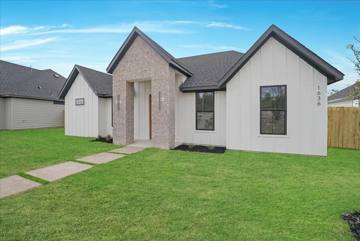 White board-and-batten siding with dark trim, brick facade entry with recessed lighting, dark shingle roof, black-framed windows, and a segmented concrete pathway