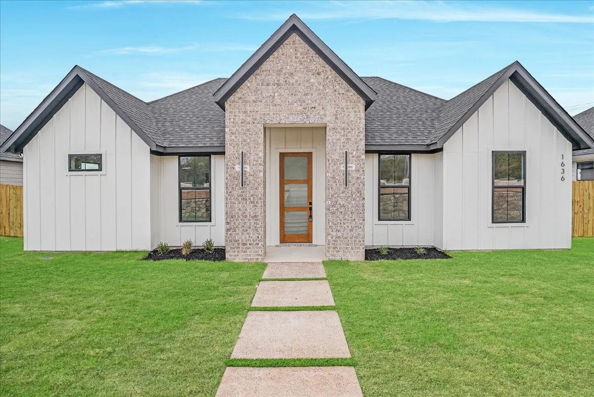 Modern farmhouse-style exterior featuring board and batten siding, a brick facade accent, dark framed windows, a wood-finish front door, and a concrete paver walkway