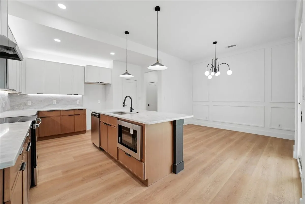 Wood-finish flooring, white wood paneling, modern chandelier, recessed lighting, and a kitchen island with built-in stainless steel appliances