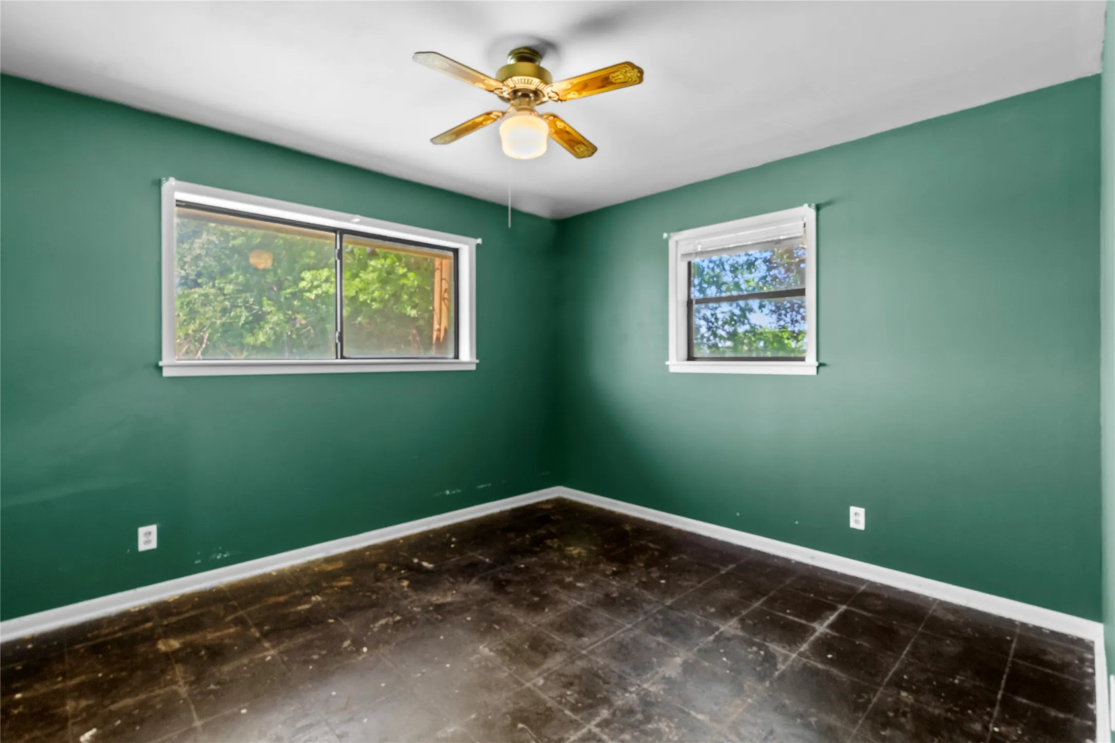Bright corner bedroom showcasing dual windows with natural light, sage green walls with crisp white trim, polished dark-finish flooring, and a brass ceiling fan with integrated light fixture