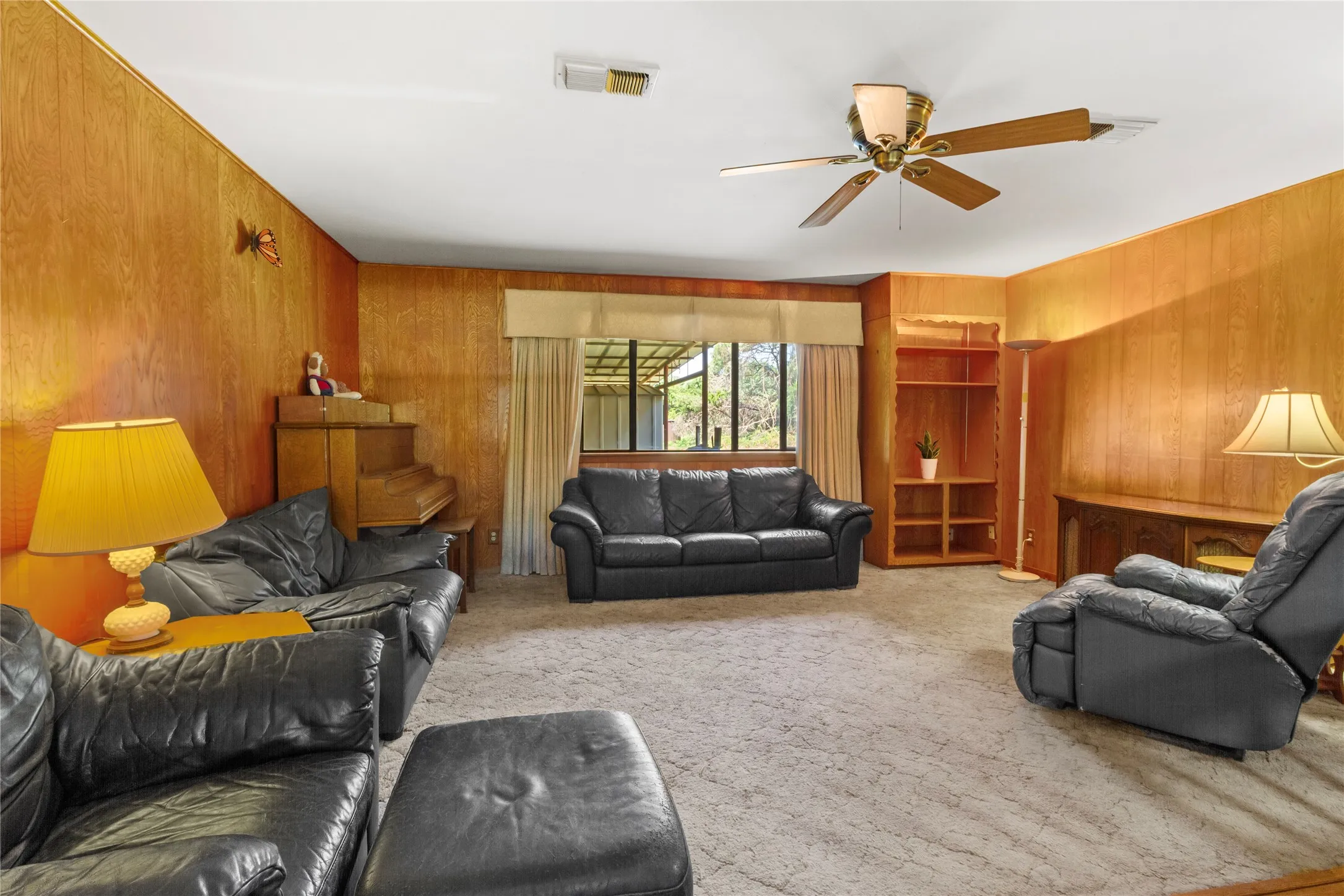 Spacious living room showcasing warm wood-paneled walls, multiple seating areas with black leather furnishings, large multi-pane windows with cream drapery, built-in wood cabinetry and shelving, ceiling fan with brass fixture, and neutral carpet flooring throughout