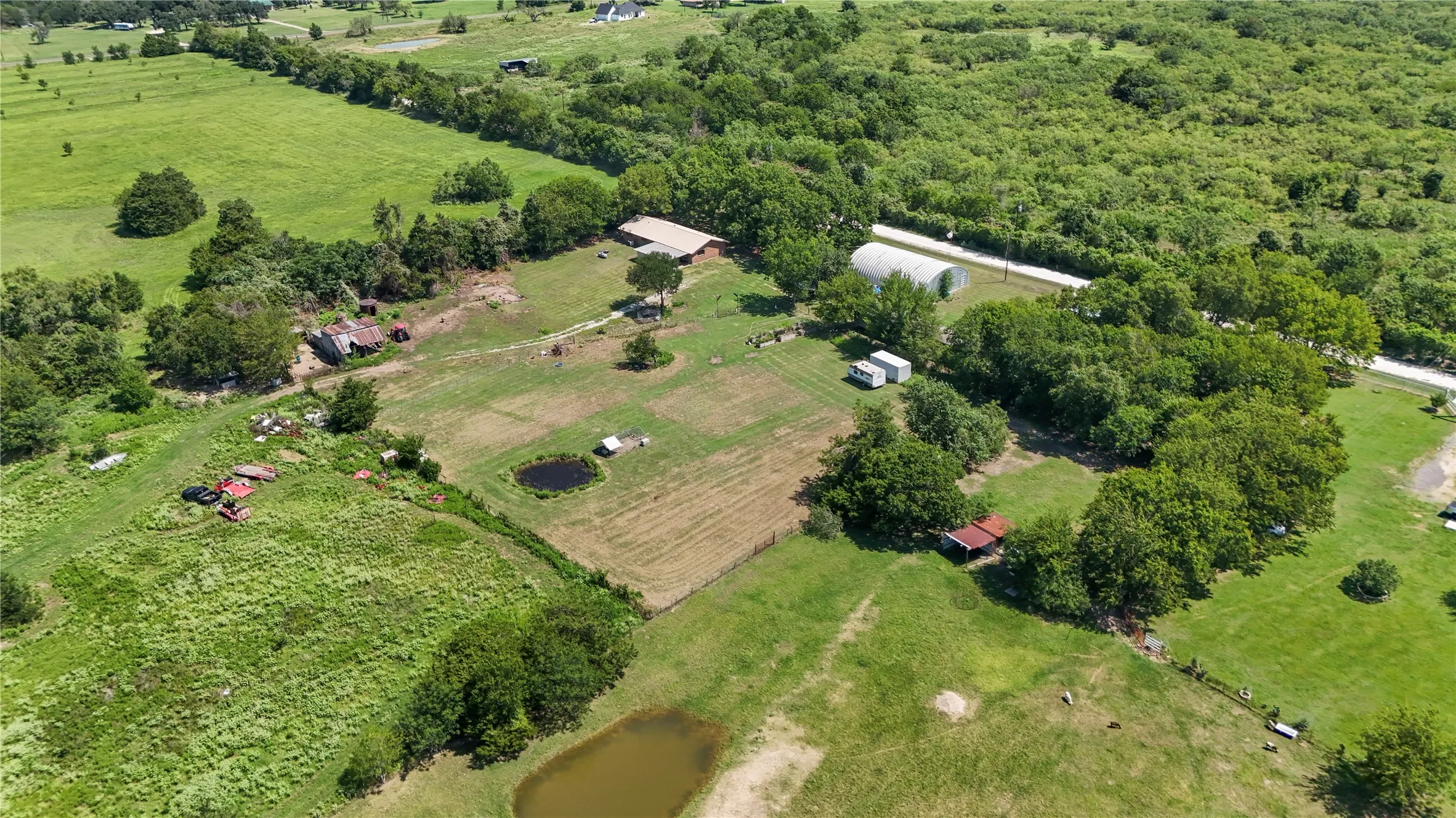 Expansive rural property featuring multiple outbuildings, mature tree-lined acreage, open pastureland, and a natural water feature