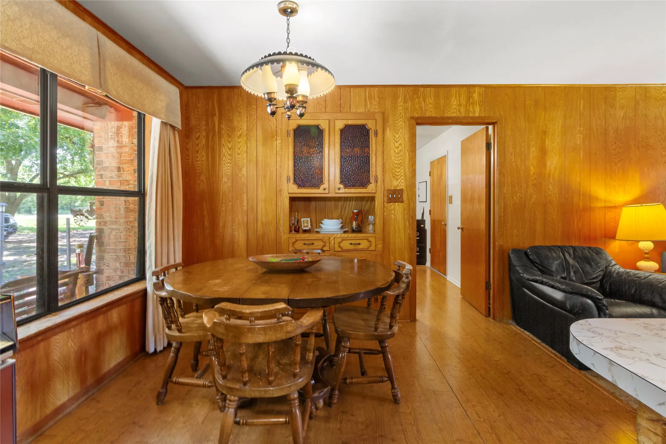 Warm wood-paneled dining room featuring a round wood dining table with coordinating chairs, wood-finish flooring throughout, and a built-in cabinet with glass-front doors