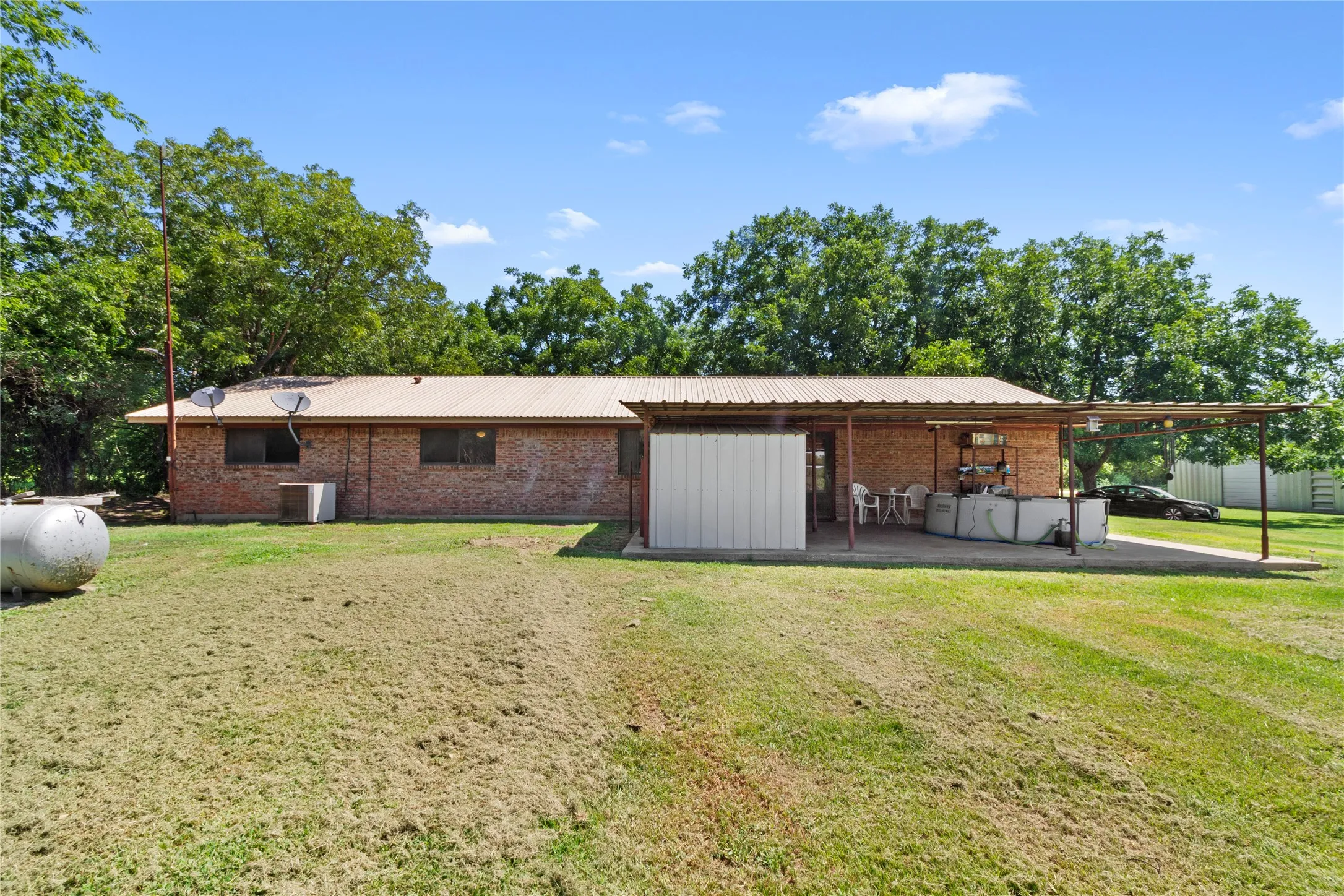 Brick residential structure with metal roof and covered carport, situated on expansive grounds with mature landscaping and established trees