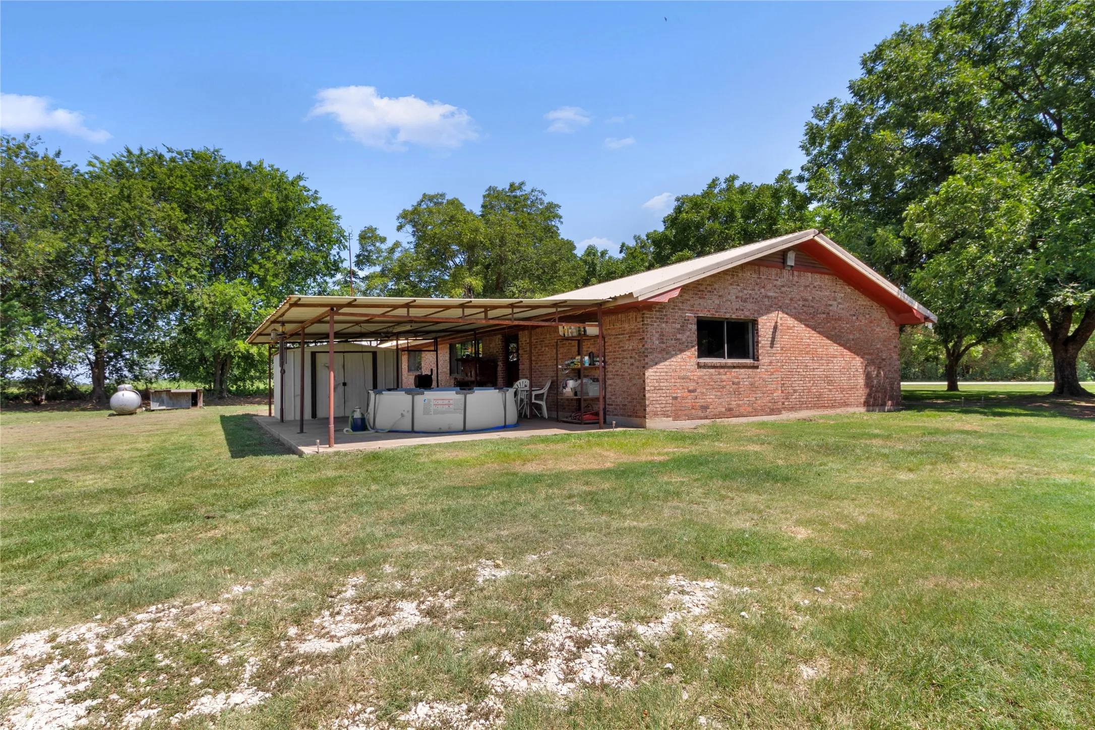 Brick-exterior structure featuring a covered patio, a small window, a pitched roof, and surrounding mature trees