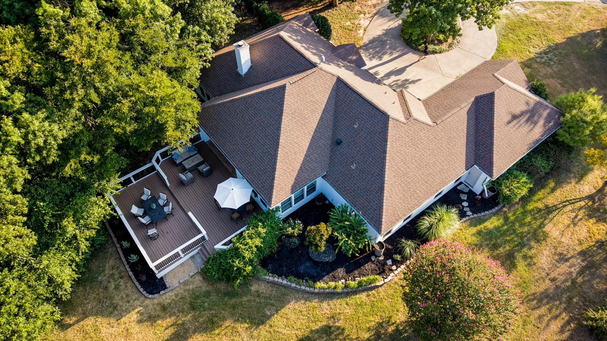 Expansive property featuring a multi-level deck, surrounding mature trees, a paved circular driveway, and detailed landscaping