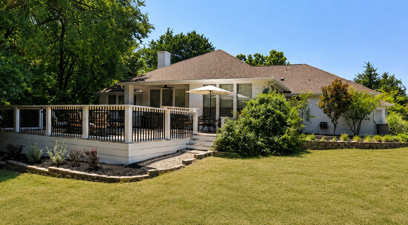 Charming single-story residence featuring a covered front porch with white balustraded railings, expansive manicured lawn, mature landscaping with established trees and shrubs, composite or wood-finish exterior siding, and a hip roof with architectural shingles