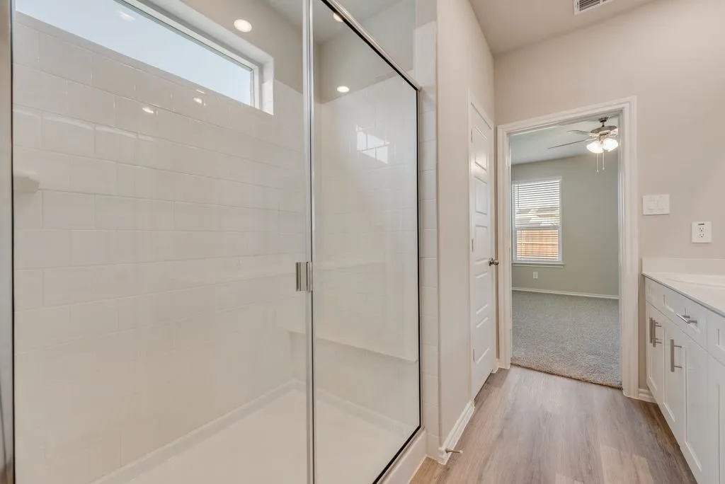 Spa-inspired primary bathroom featuring a frameless glass-enclosed walk-in shower with subway tile surround, dual vanity with white cabinetry and integrated lighting, wood-finish flooring, and a separate water closet with natural light from transom window