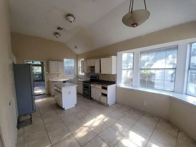 Bright, sun-filled kitchen featuring white cabinetry, tile flooring, multiple large windows with natural light, a central island with prep space, and stainless steel appliances