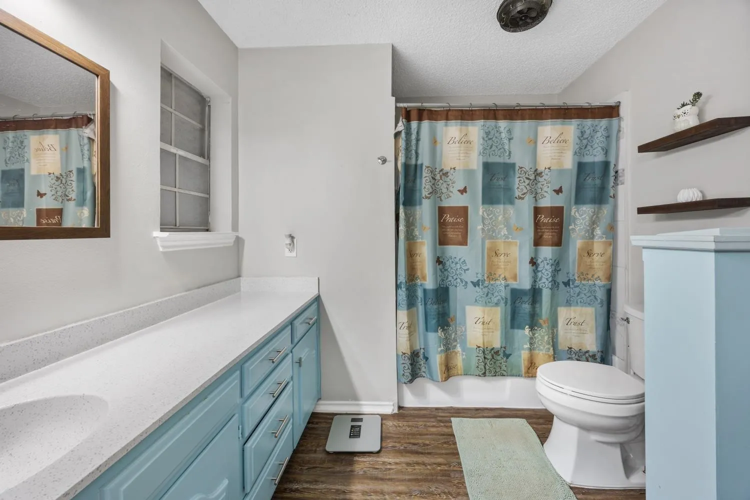 Bathroom featuring wood-finish flooring, a light blue vanity with a white countertop and integrated sink, a framed mirror, a window with blinds, and a built-in shower/tub combination