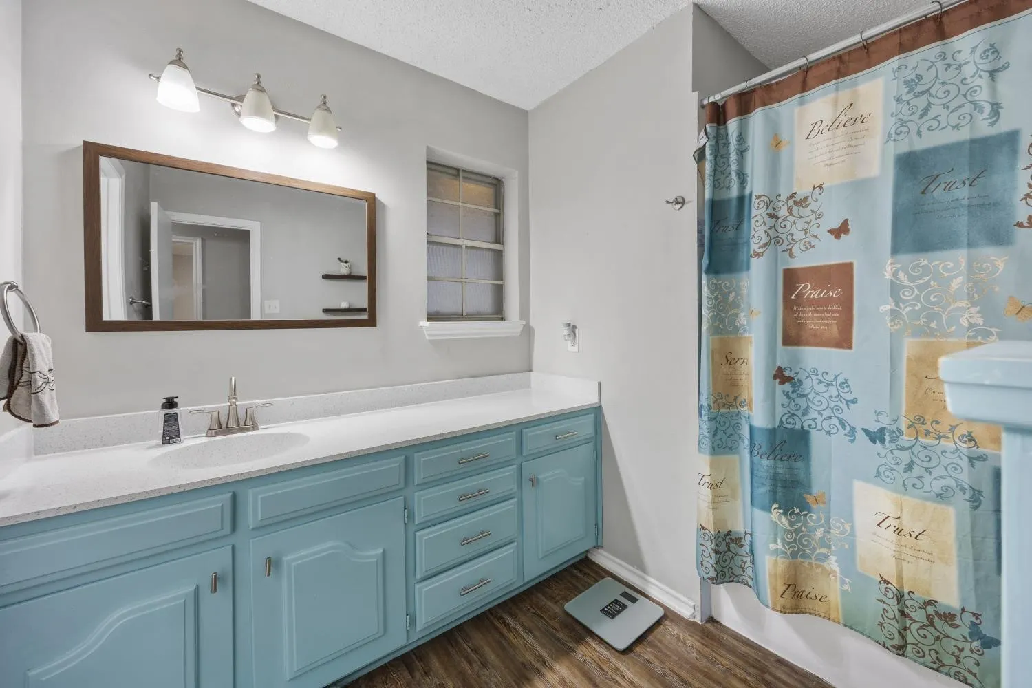 Spa-inspired primary bathroom featuring a turquoise vanity with multiple drawers and cabinet storage, white quartz countertop, brushed nickel fixtures, and an illuminated rectangular mirror