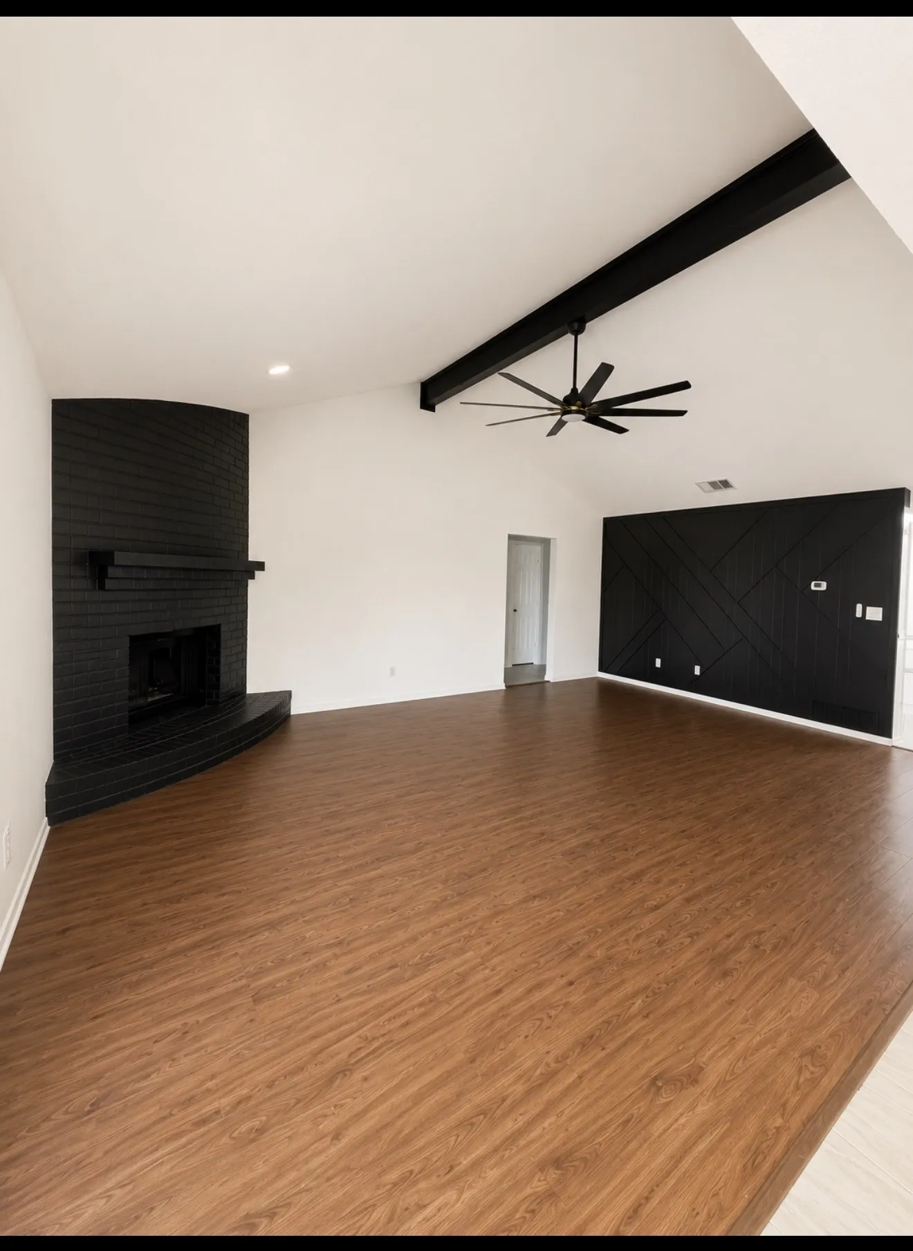 Spacious living area showcasing vaulted ceilings with exposed black beam detail, wood-finish flooring throughout, a black brick fireplace with linear design, and a ceiling fan with integrated lighting