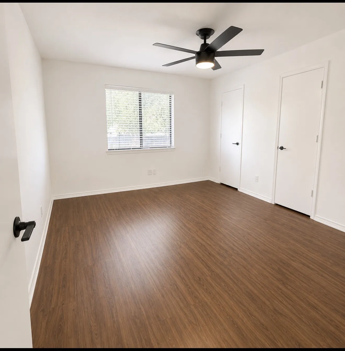 Spacious bedroom showcasing wood-finish flooring, ceiling fan with integrated lighting, dual closets with white panel doors, and large window with blinds providing natural light