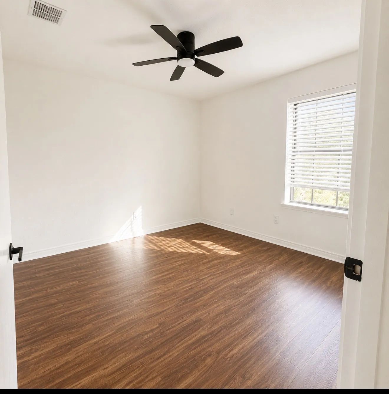 Bright and spacious bedroom featuring warm wood-finish flooring, ceiling fan with integrated lighting, and abundant natural light from large window with plantation shutters