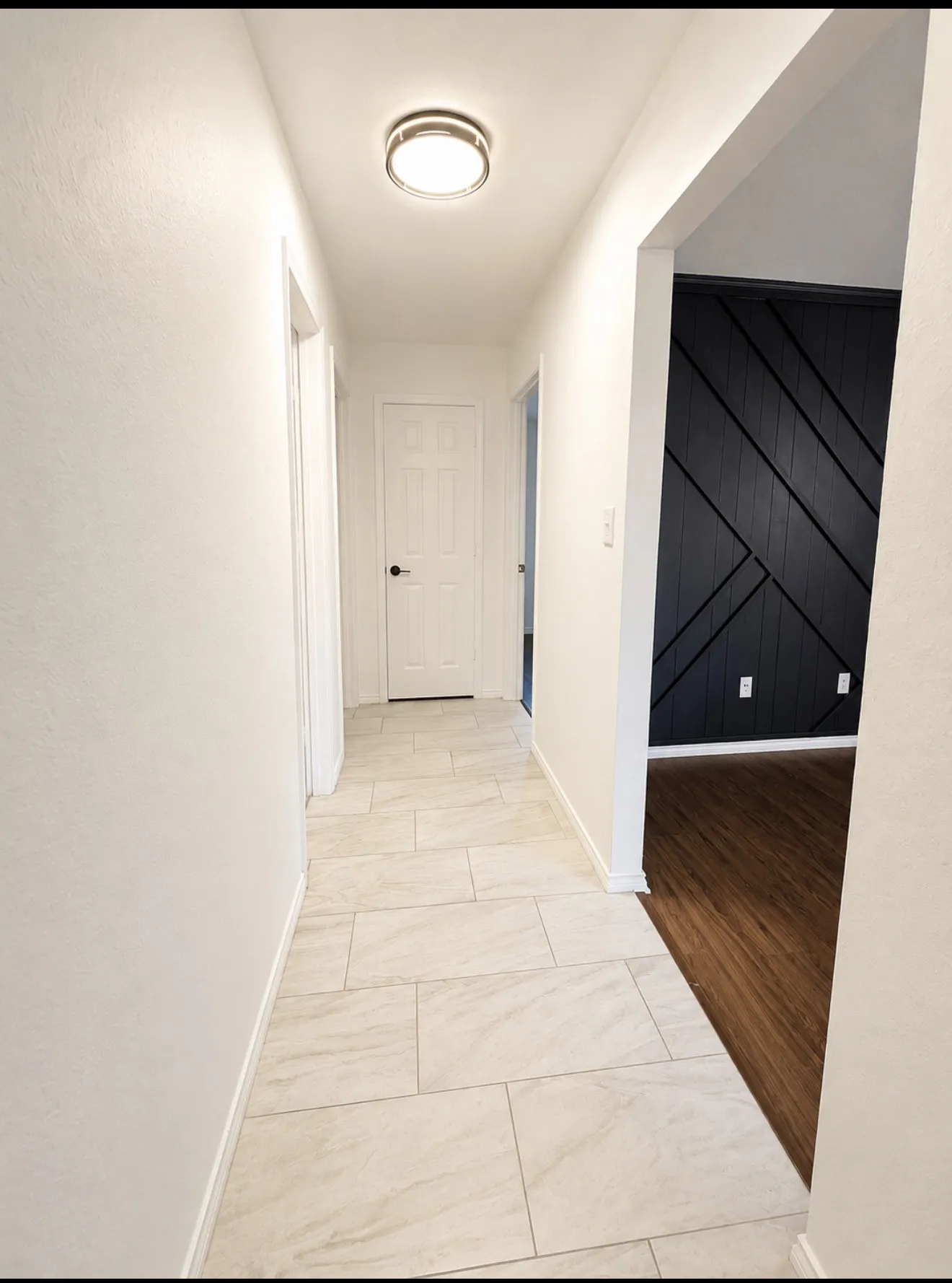Modern hallway featuring light-toned tile flooring with linear layout, recessed ceiling lighting, crisp white walls, and direct access to multiple rooms including a bedroom with black shiplap accent wall and wood-finish flooring