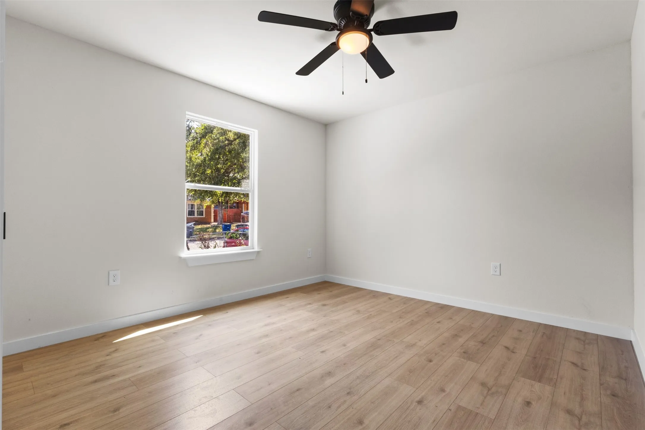 Bright and spacious bedroom featuring hardwood-finish flooring, a ceiling fan with integrated lighting, and abundant natural light from a large window framing outdoor scenery