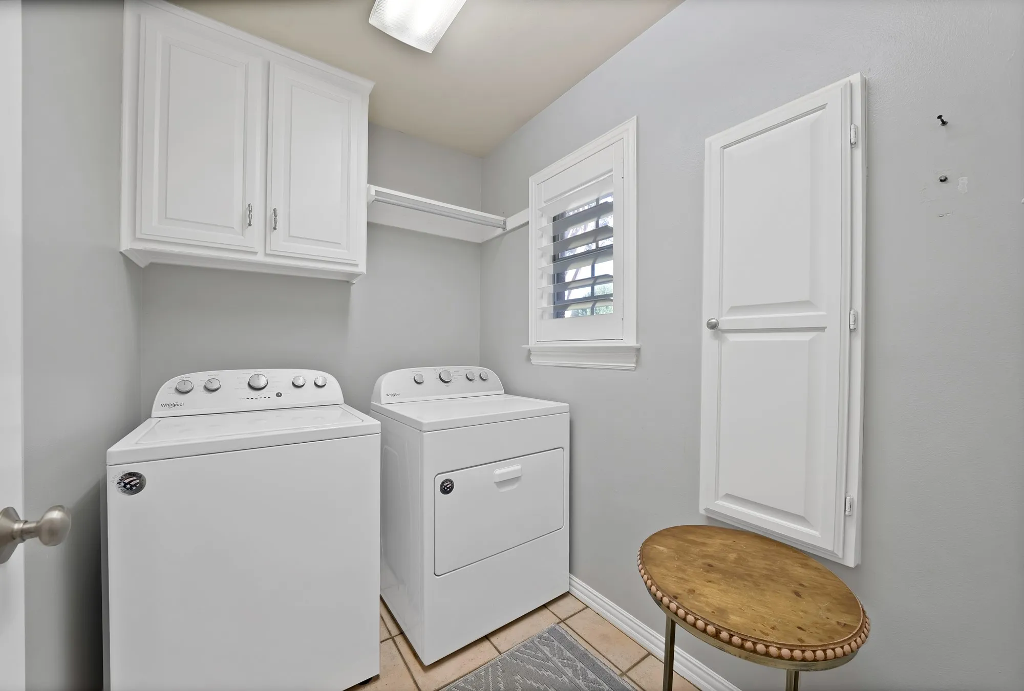 Utility room featuring overhead cabinetry, wall-mounted shelving, tiled flooring, and a window with plantation shutters