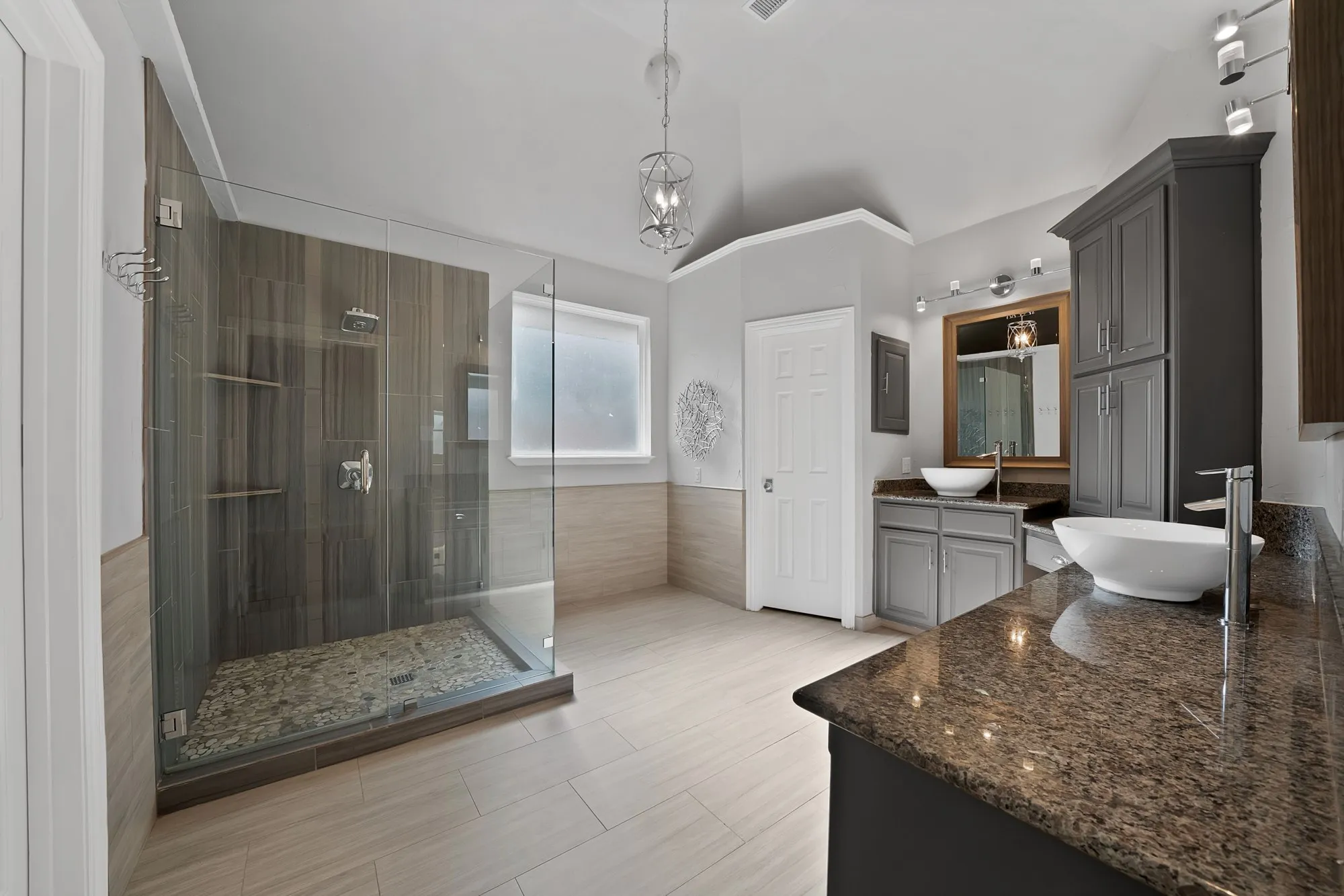 Spacious bathroom featuring a glass-enclosed shower with pebble flooring and wood-look tile surround, dual vanity with vessel sinks, granite countertops, and light wood-finish flooring