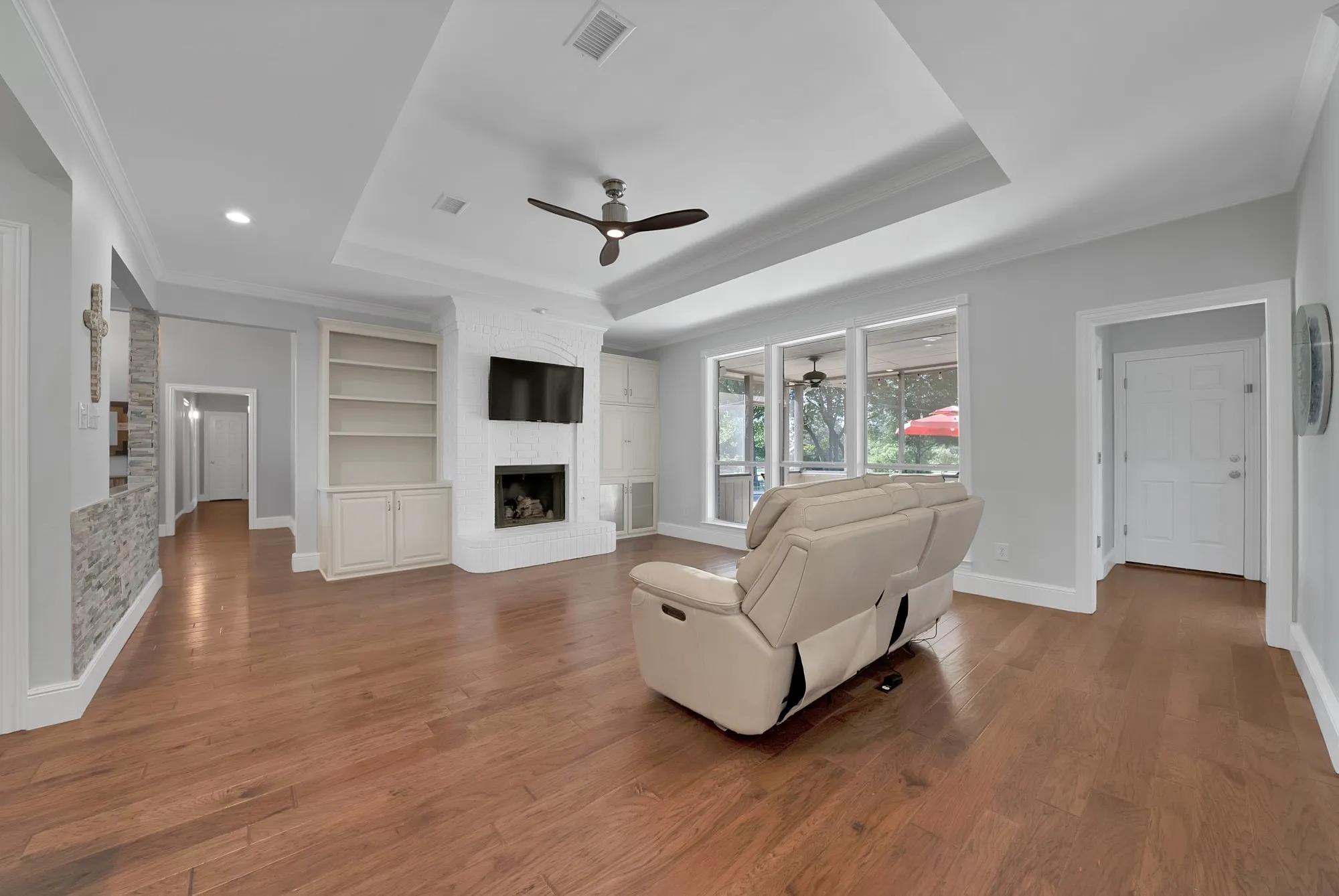 Expansive living area featuring wood-finish flooring, a white brick fireplace with built-in cabinetry, a tray ceiling, and a ceiling fan