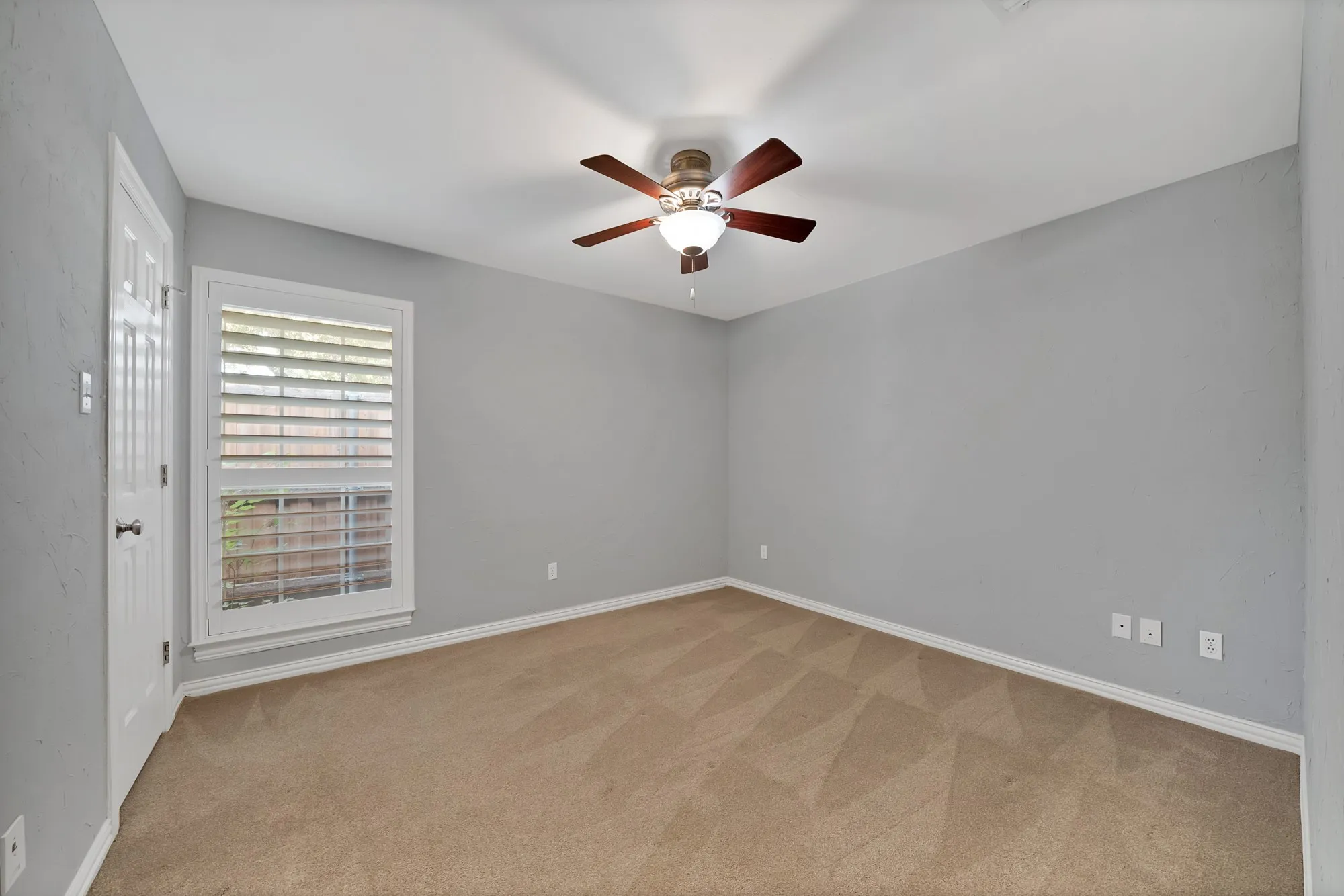 Spacious bedroom featuring neutral gray walls, wood-finish flooring, and abundant natural light from a large window with plantation shutters