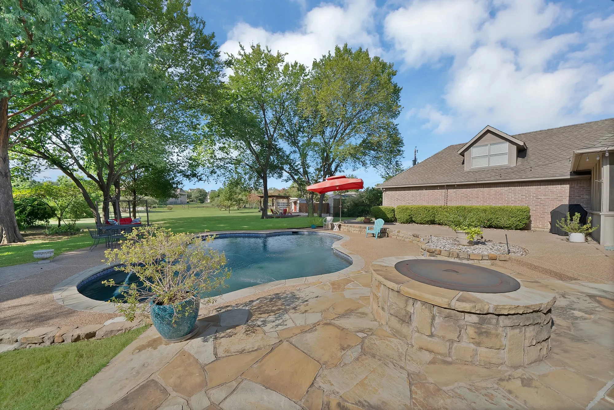 Freeform swimming pool with flagstone surround, a stone fire pit, and extensive hardscaping, complemented by a large grassy area and mature trees