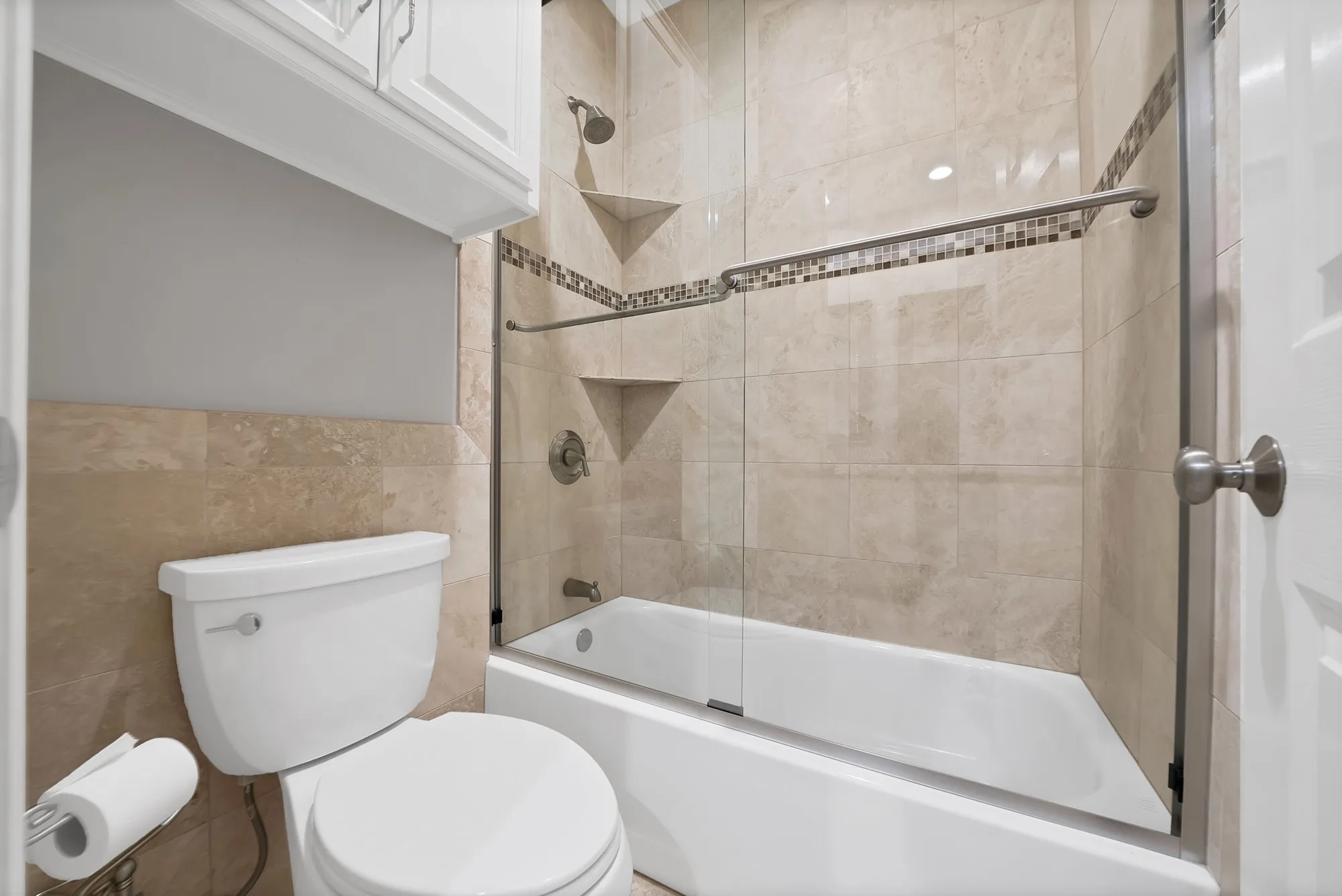 Updated bathroom featuring ceramic tile walls with decorative border accent, white porcelain toilet, and a frameless glass-enclosed shower with white subway tile surround and dual shelving niches