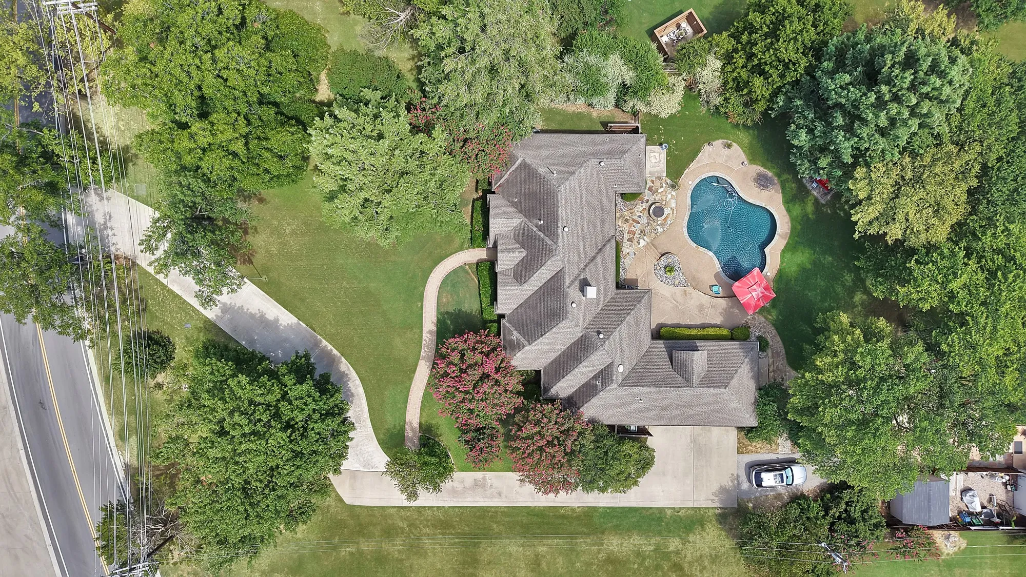 Aerial view showcasing this residence's expansive grounds featuring a resort-style swimming pool with curved design, manicured lawn, mature tree-lined perimeter, circular driveway, and multi-level architectural design with gray tile roofing