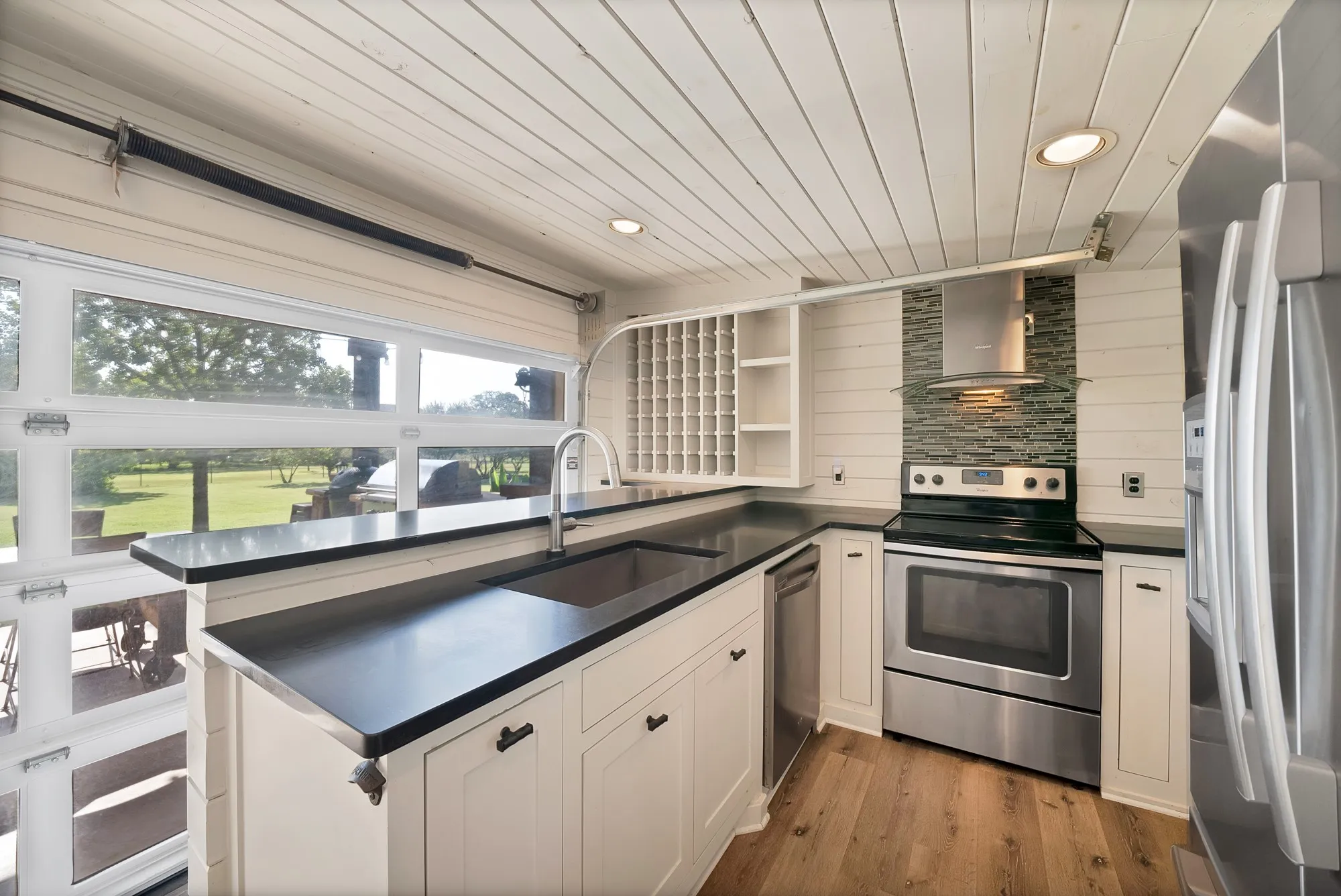Kitchen featuring a roll-up glass panel door, white shiplap walls, wood-finish flooring, dark countertops, and stainless steel appliances