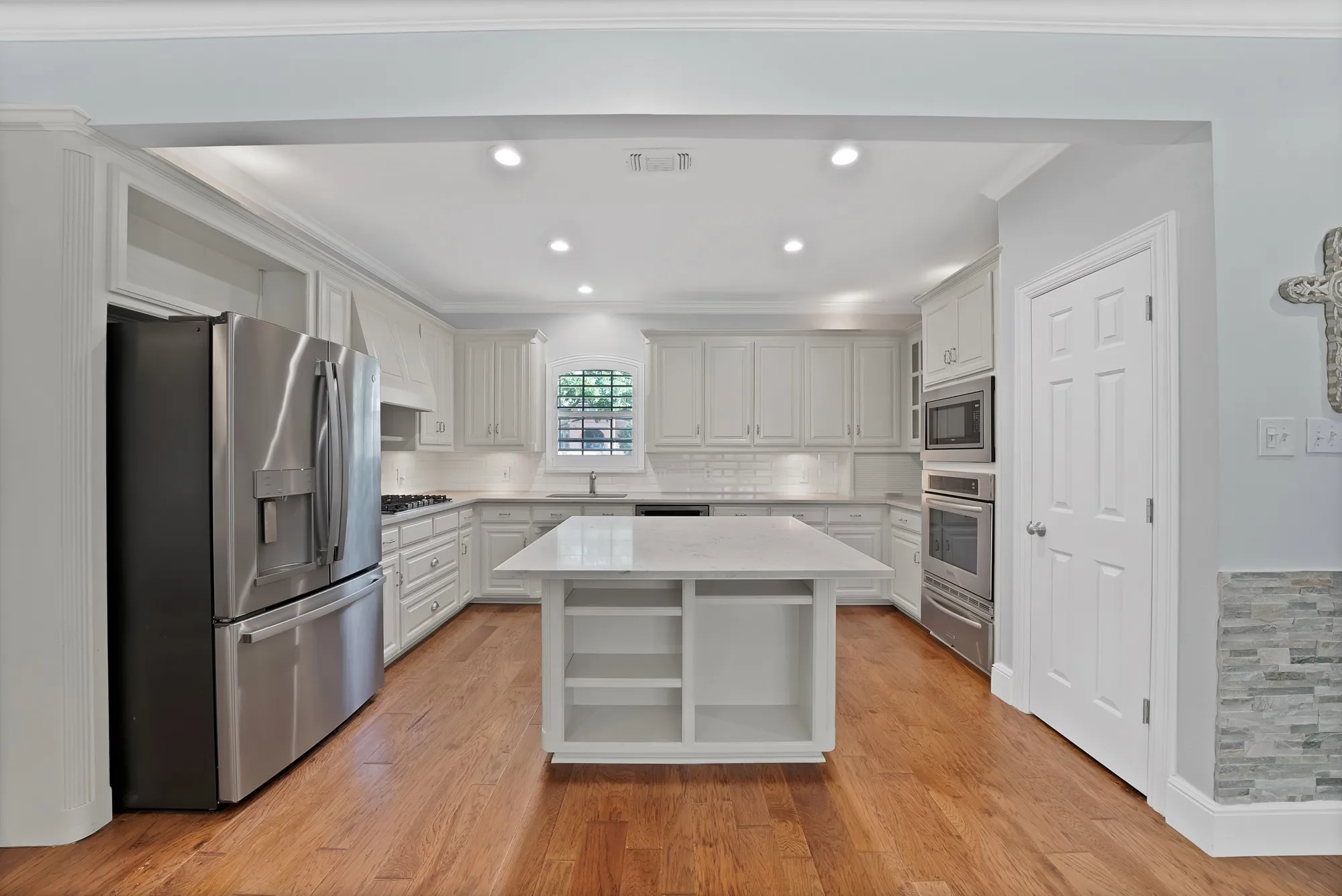 Gourmet kitchen featuring a spacious center island with open shelving, stainless steel French-door refrigerator, dual wall ovens, white cabinetry with soft-close doors, quartz countertops, recessed lighting, and warm wood-finish flooring throughout