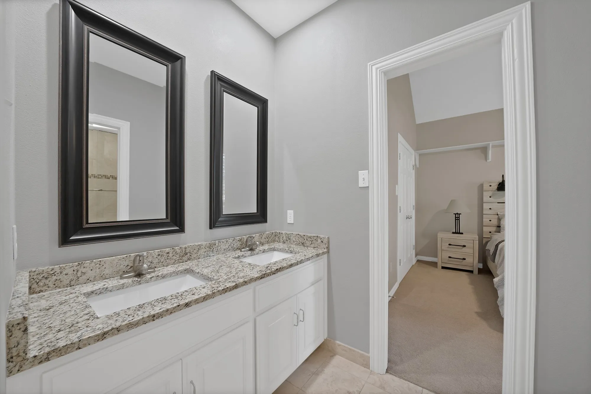 Dual vanity bathroom featuring a granite-topped countertop with two rectangular sinks, framed black-trimmed mirrors, white cabinetry with hardware accents, and neutral tile flooring