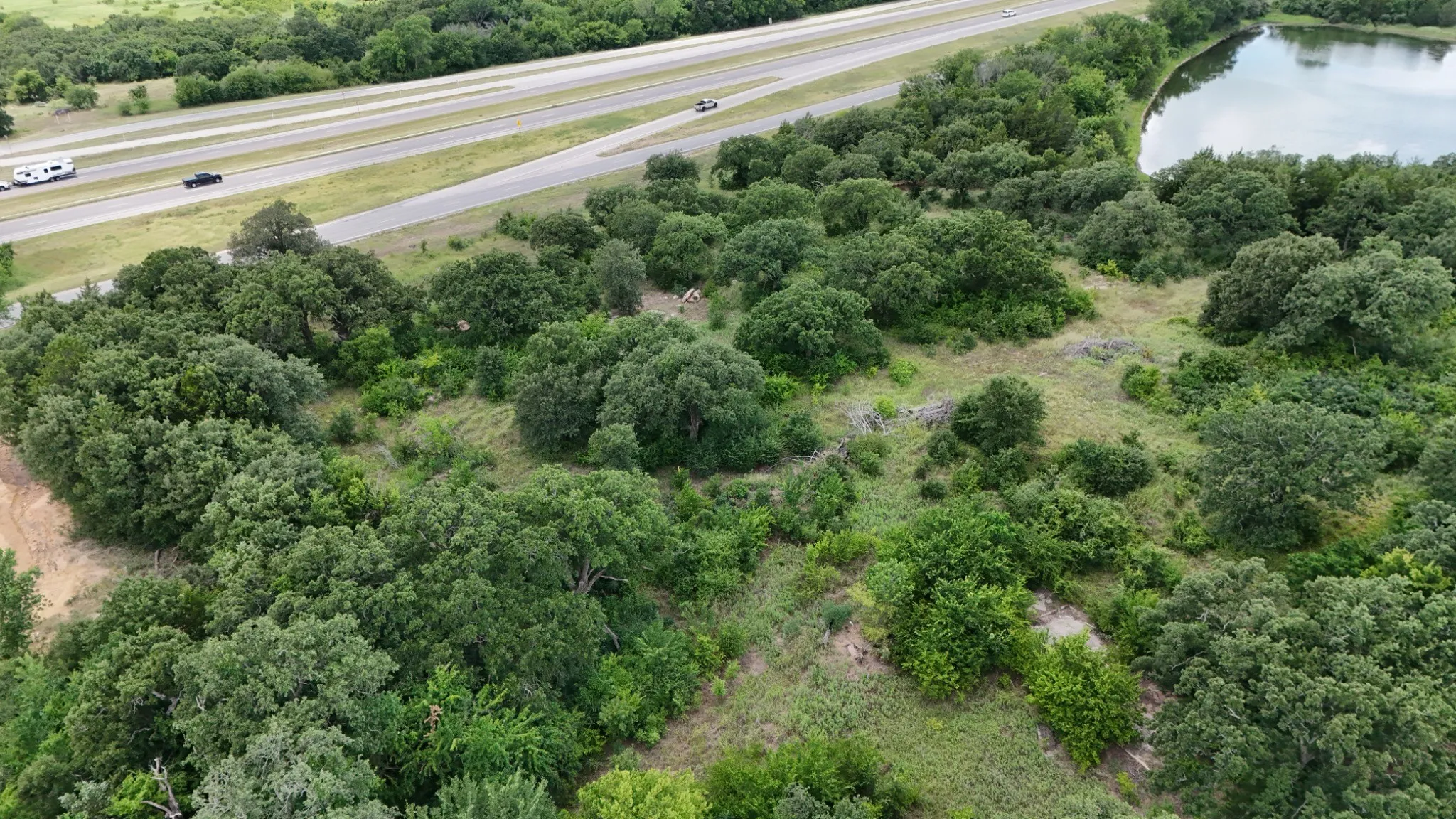 Expansive wooded acreage featuring mature hardwood and oak trees with natural ground cover, adjoining highway access and scenic water feature visible in proximity to the property