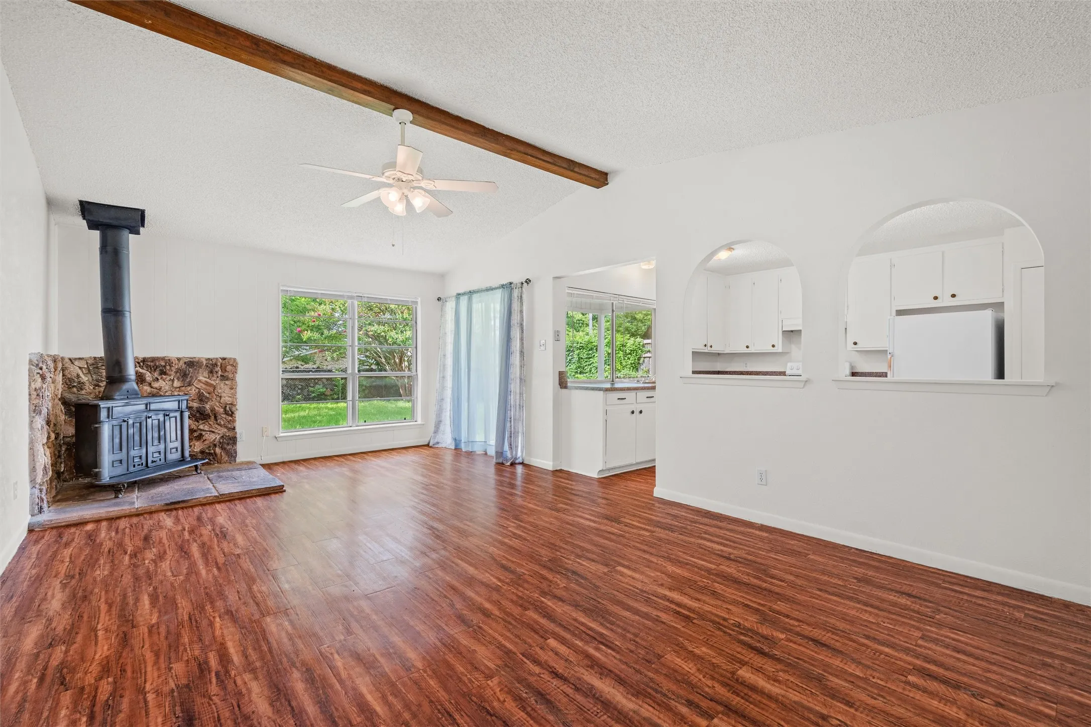 Warm wood-look flooring, a vaulted ceiling, exposed beam, and dramatic stone-backed wood-burning stove create a living space filled with character. Large windows bring the outdoors in, while arched openings keep the layout open and inviting.