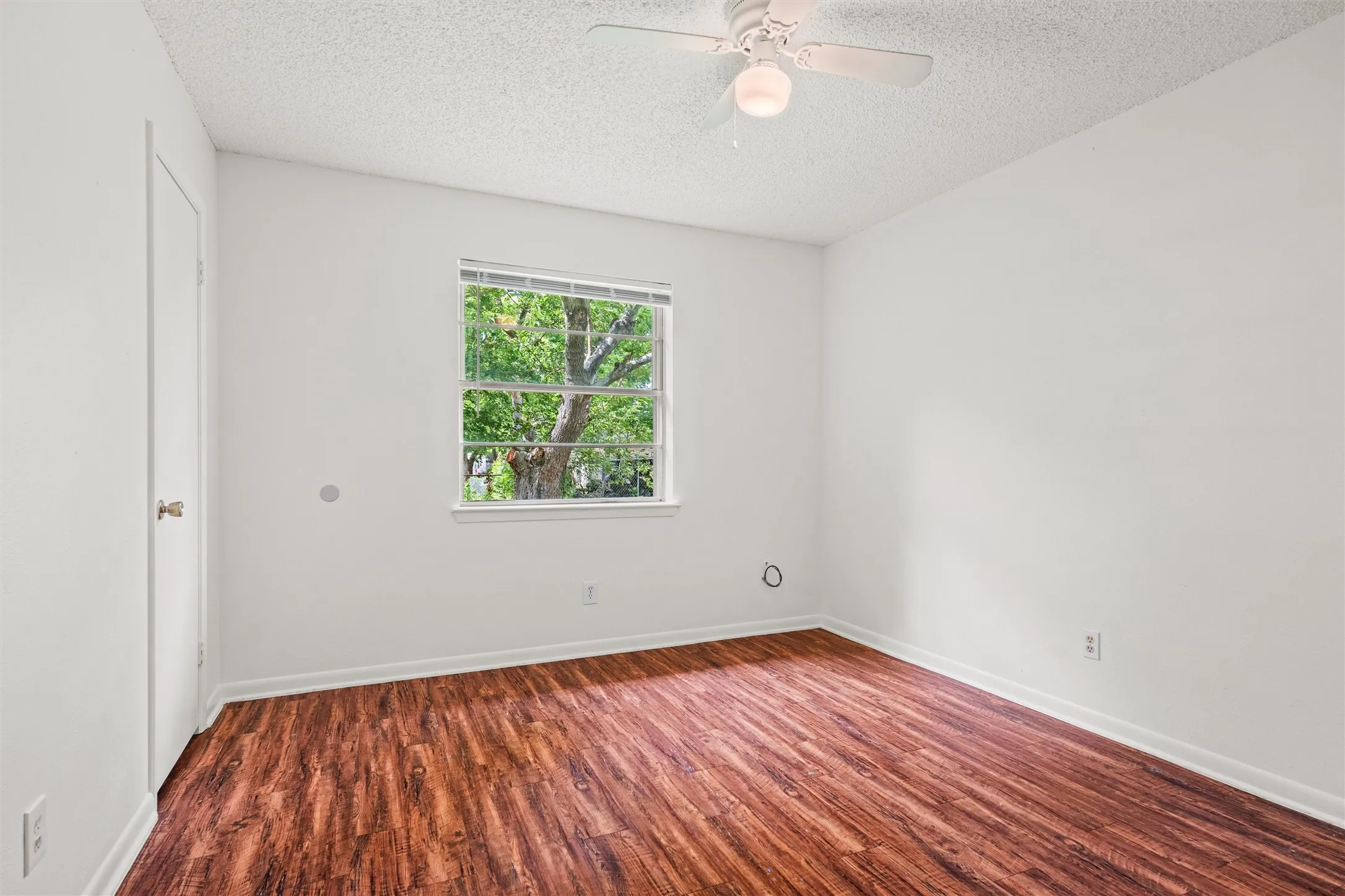 A bright bedroom with warm flooring, a ceiling fan, closet storage, and a calming view of the mature trees outside creates the perfect place to relax and recharge.