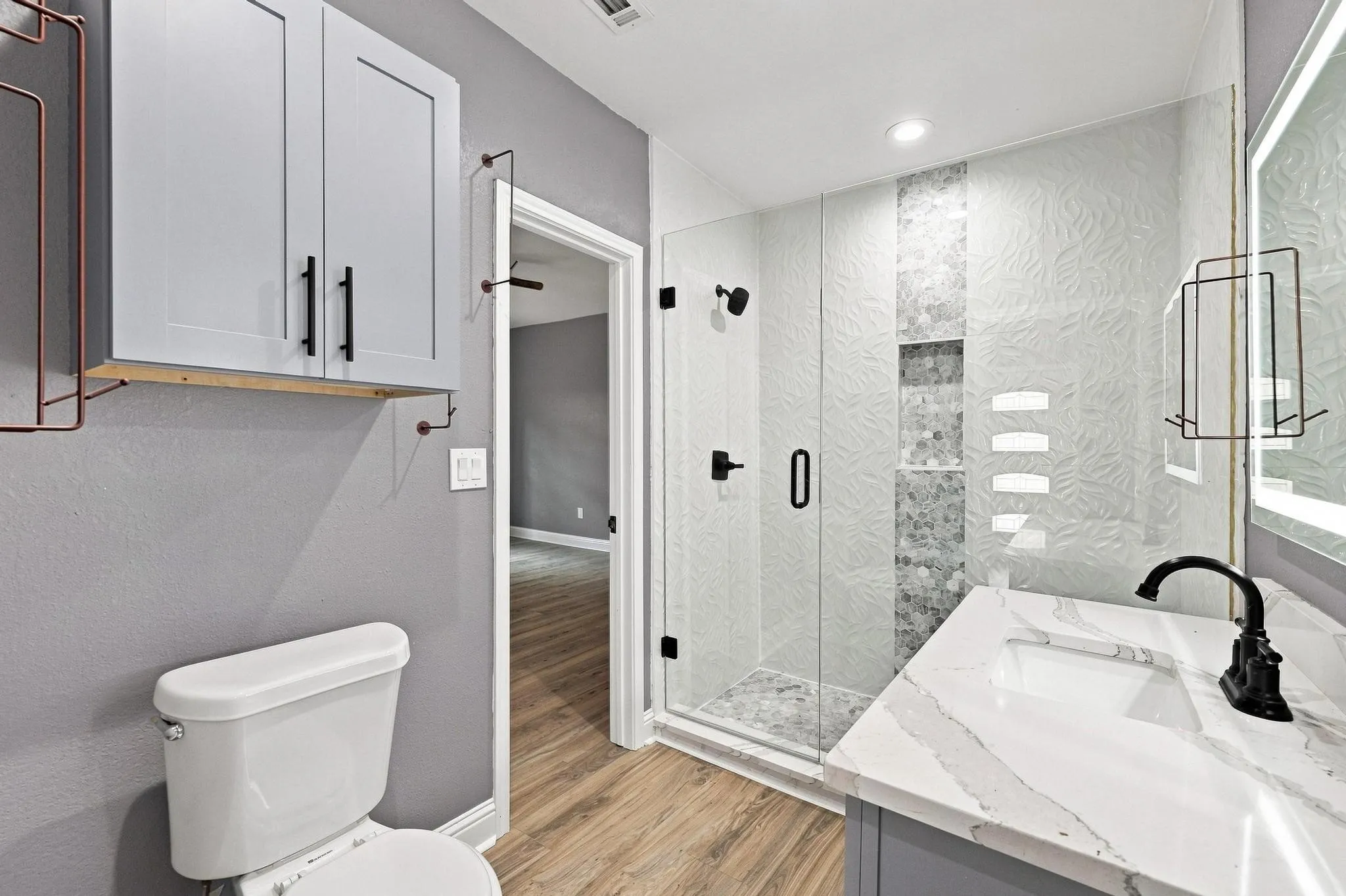 Glass-enclosed shower with textured tile, vanity with integrated sink and stone countertop, wood-finish flooring, and wall-mounted cabinetry
