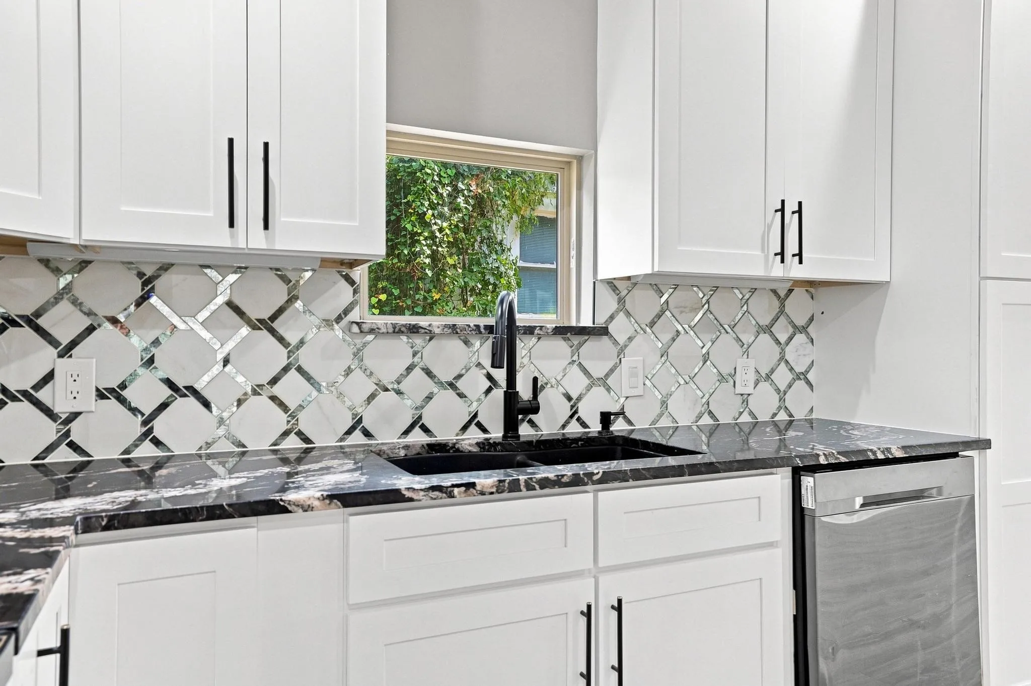 White cabinetry with matte black hardware, dark stone-finish countertops, black sink and faucet, patterned tile backsplash, and a stainless steel dishwasher