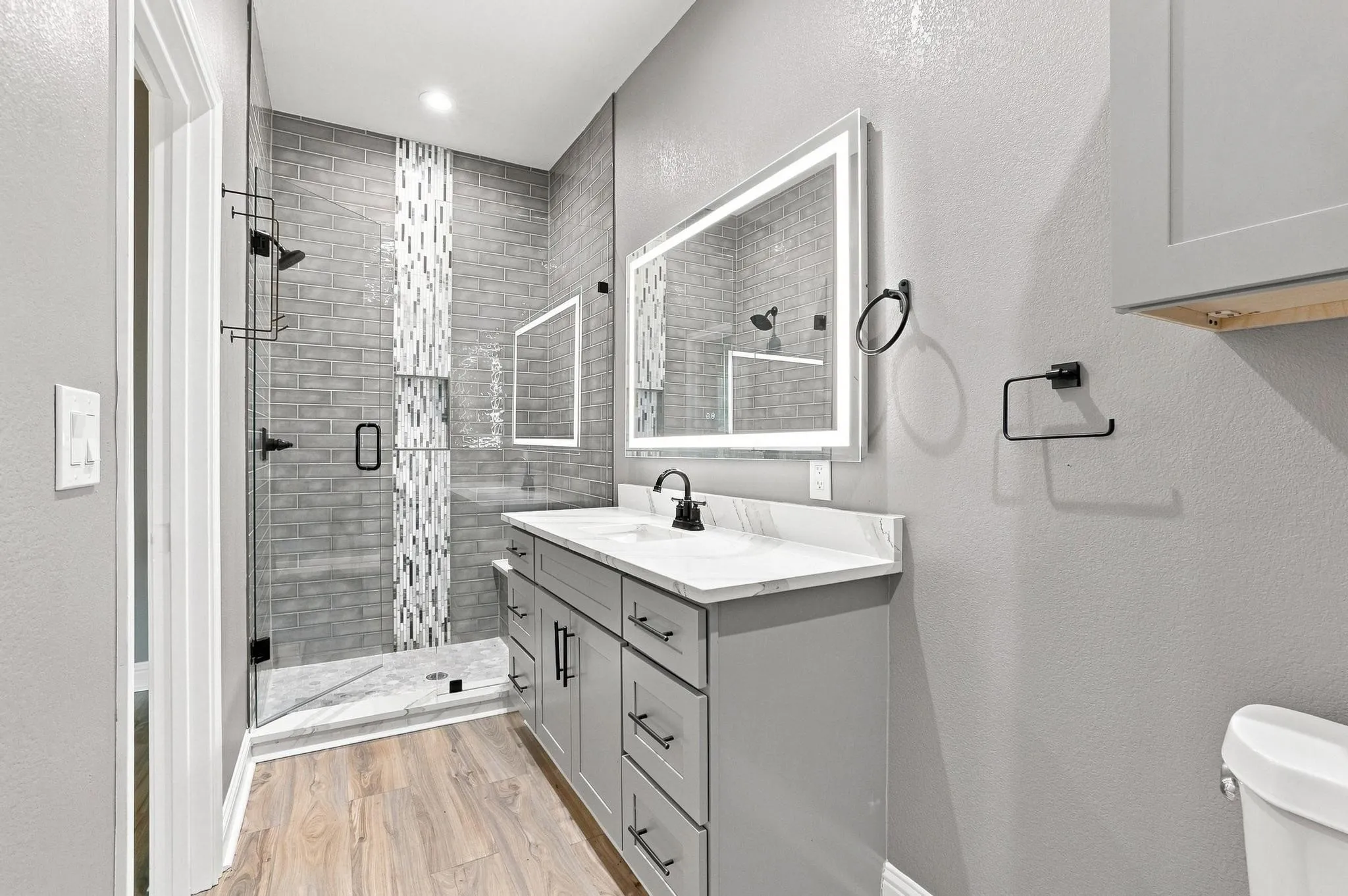 Walk-in shower with subway tile and glass enclosure, dual vanity with quartz countertop, light wood-finish flooring, and illuminated mirror