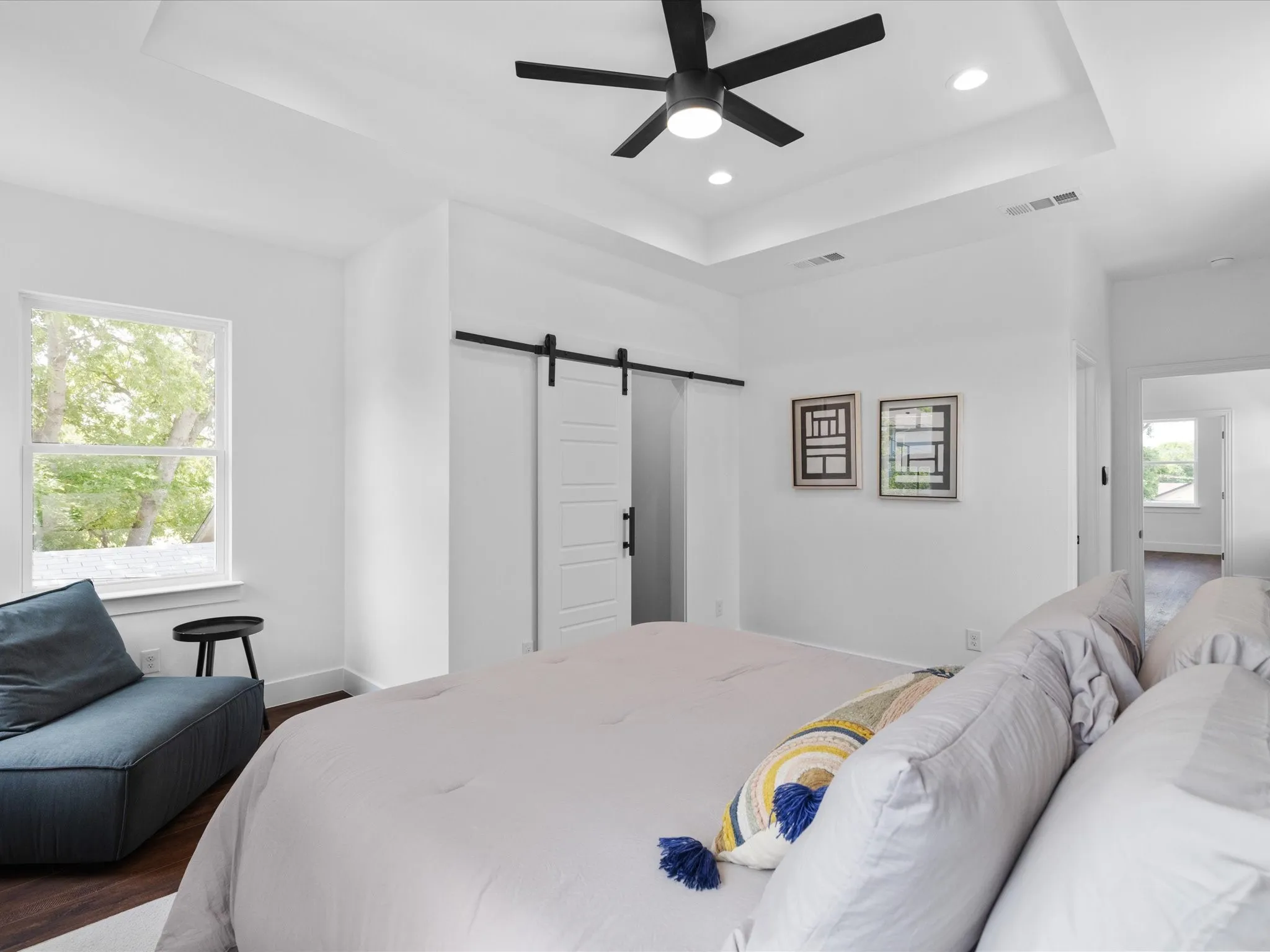 Spacious room featuring a tray ceiling with recessed lighting and a black ceiling fan, wood-finish flooring, a contemporary barn door, and a large window