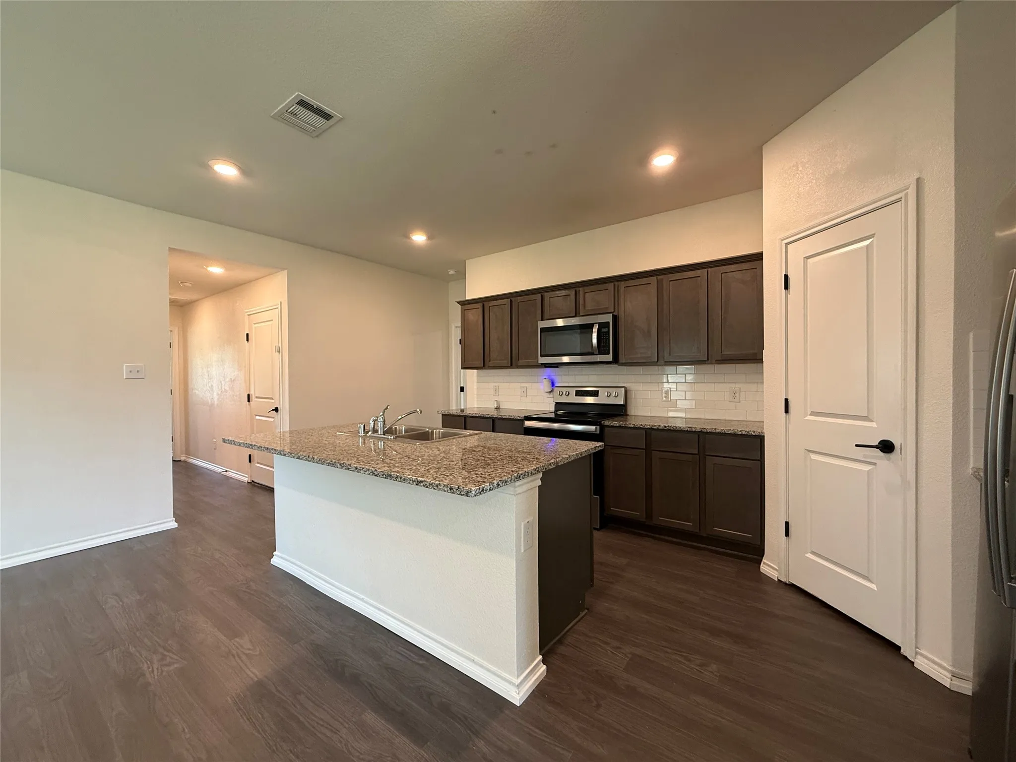Kitchen featuring an island with a built-in sink, granite countertops, wood-finish flooring, dark wood cabinetry, and stainless steel appliances