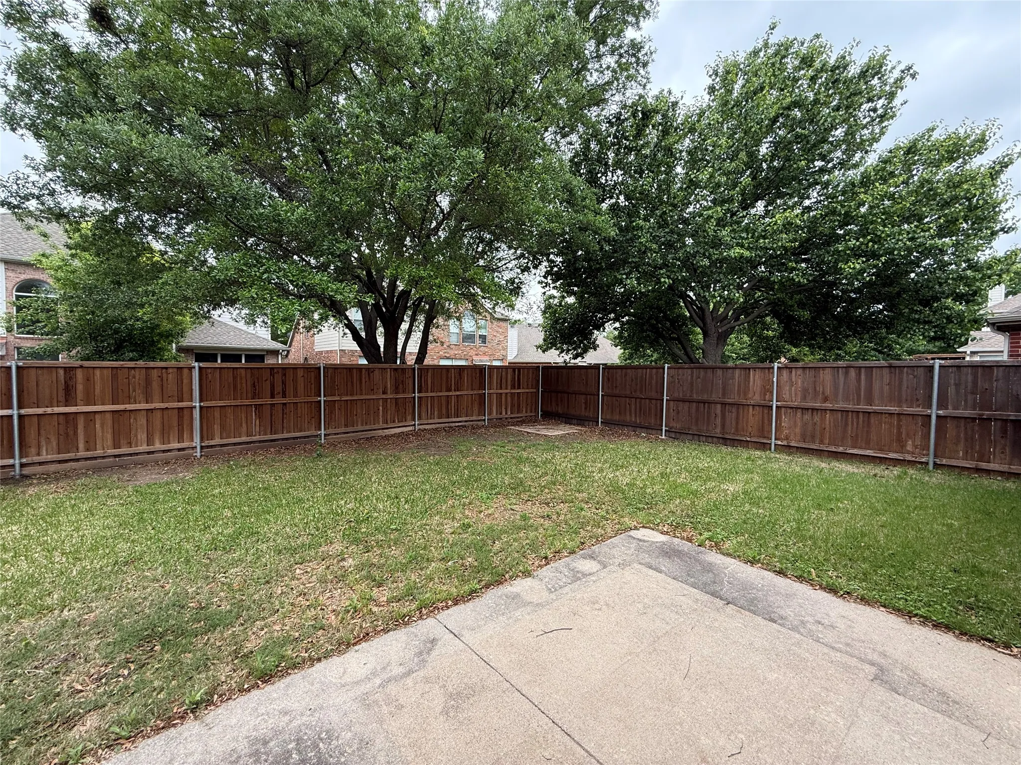 Expansive backyard featuring a wood privacy fence, mature trees, a concrete patio, and a grass lawn