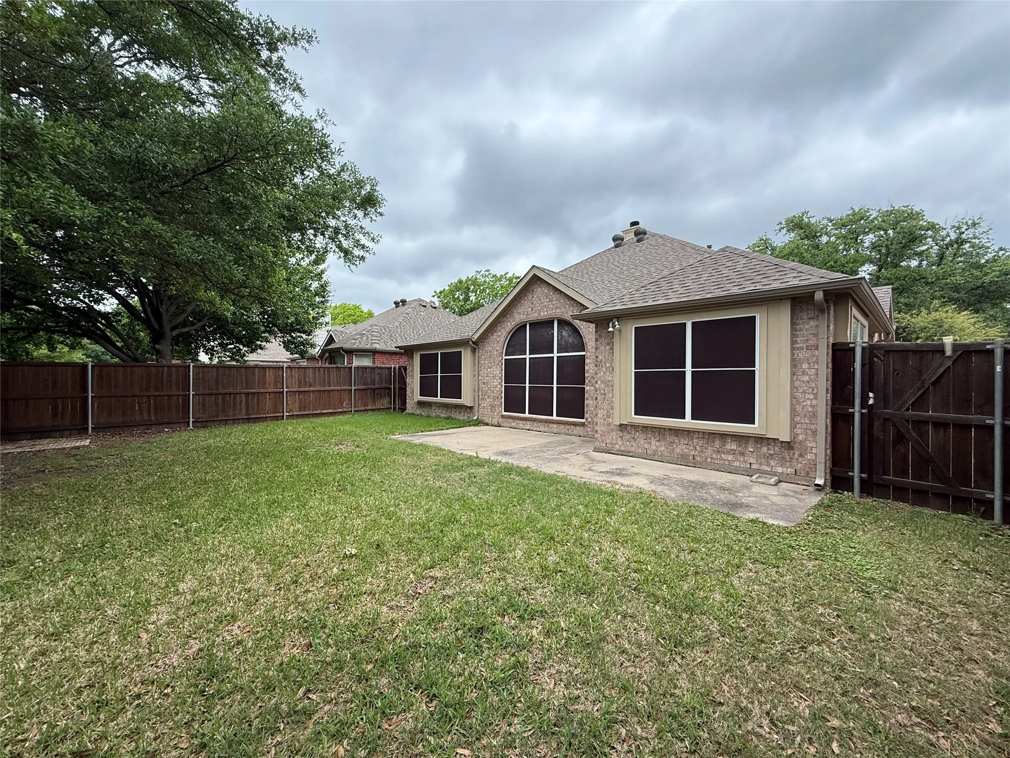 Expansive green lawn with a brown privacy fence, a concrete patio, and a brick exterior with framed windows and an arched window
