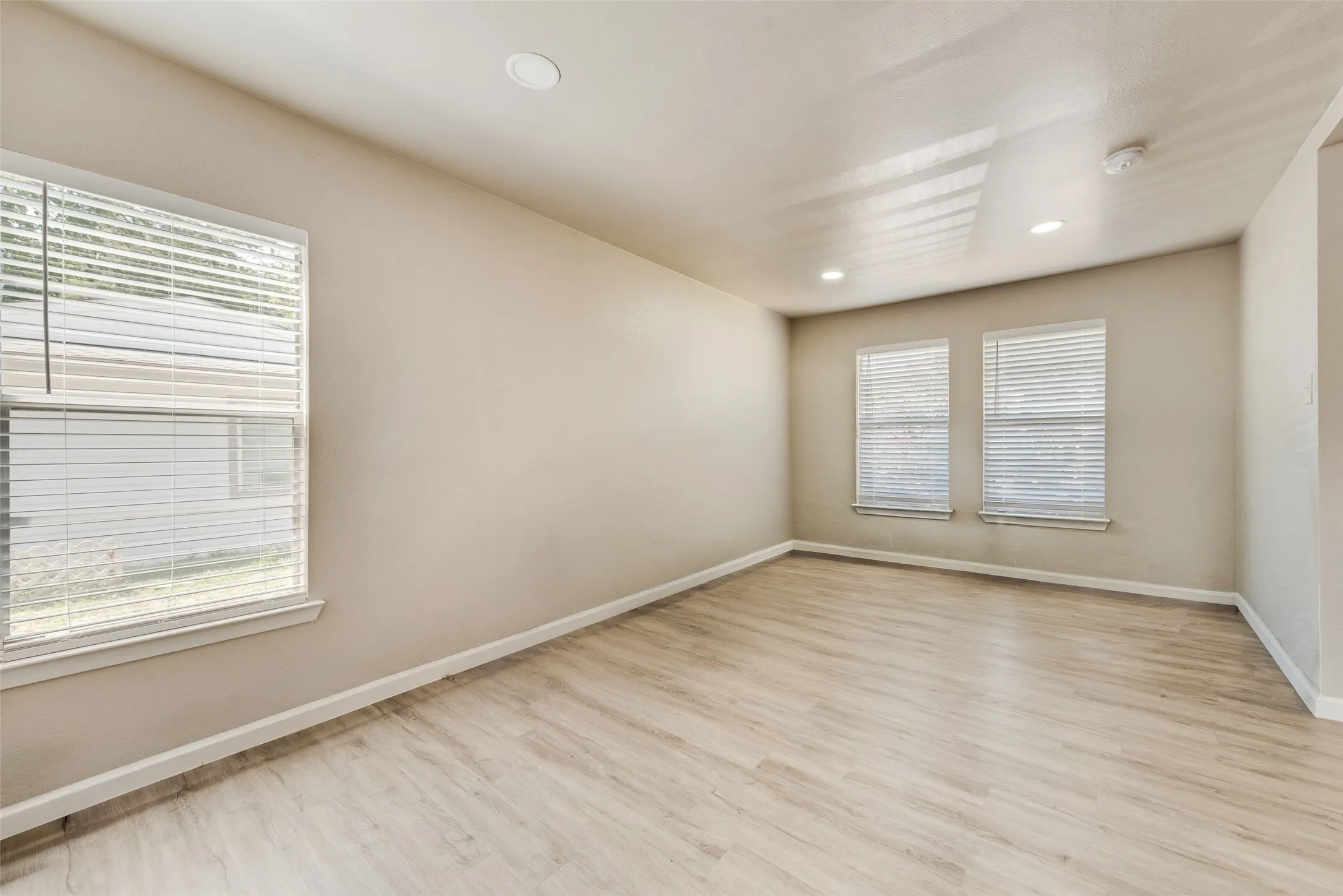 Spacious room featuring light wood-finish flooring, recessed lighting, multiple windows with blinds, and neutral-toned walls