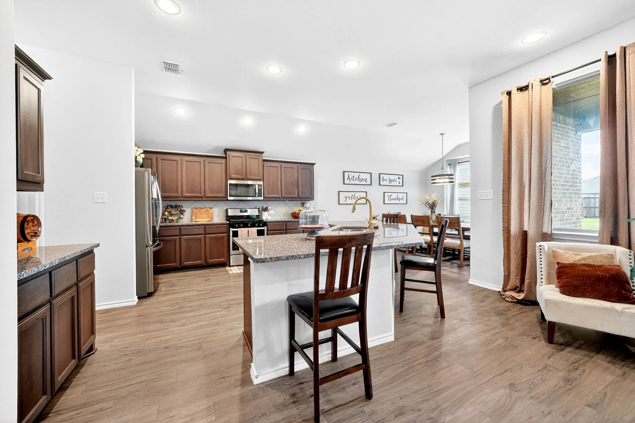 Open-concept kitchen and dining area featuring rich wood cabinetry, stainless steel appliances, a large island with seating, wood-finish flooring throughout, and abundant natural light from multiple windows and recessed ceiling lighting