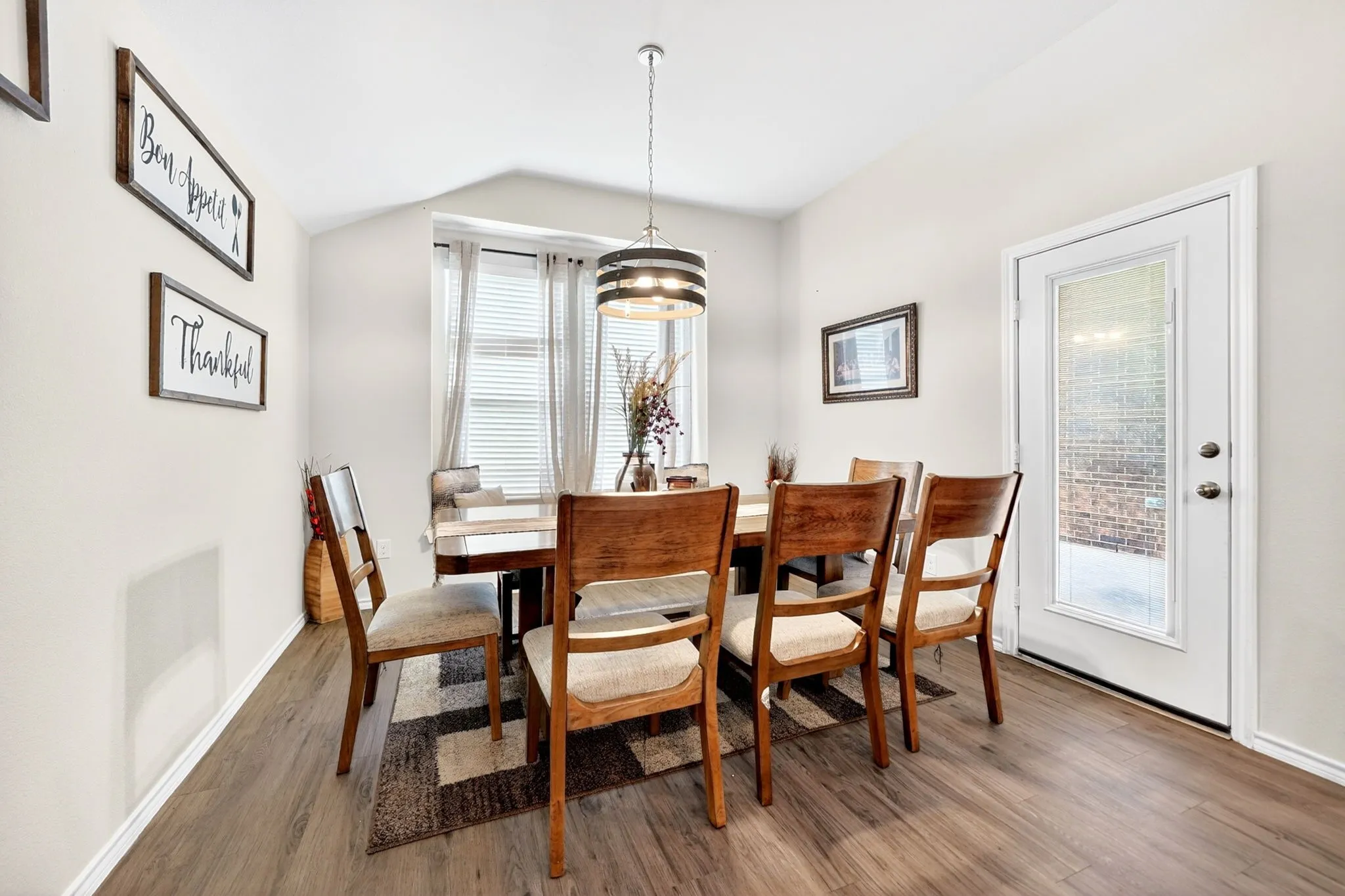 Dining area featuring durable wood-finish flooring, a contemporary chandelier, a window with blinds and sheer curtains, and a glass-paneled exterior door