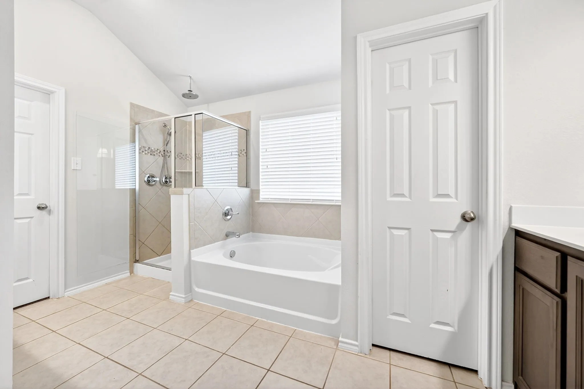 Spacious primary bathroom featuring a frameless glass-enclosed shower with tile surround, a soiled tub with integrated surround, dual vanity sinks, neutral tile flooring, recessed ceiling lighting, and abundant natural light from a large window