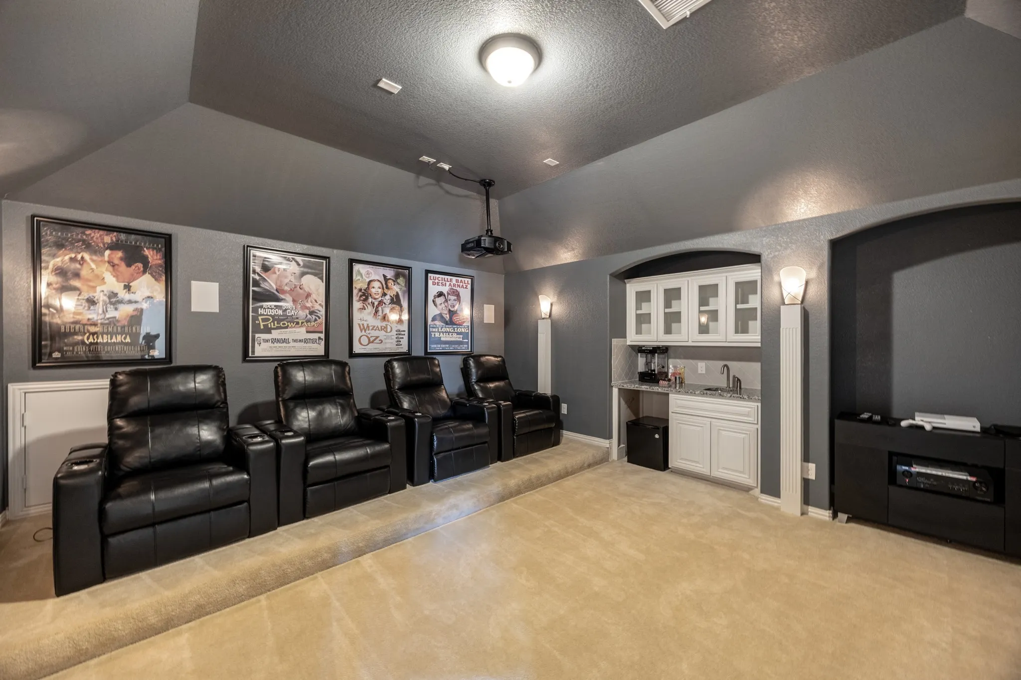 Theater room featuring a tiered carpeted floor, recessed projector, and integrated wet bar with white cabinetry and a light-toned countertop