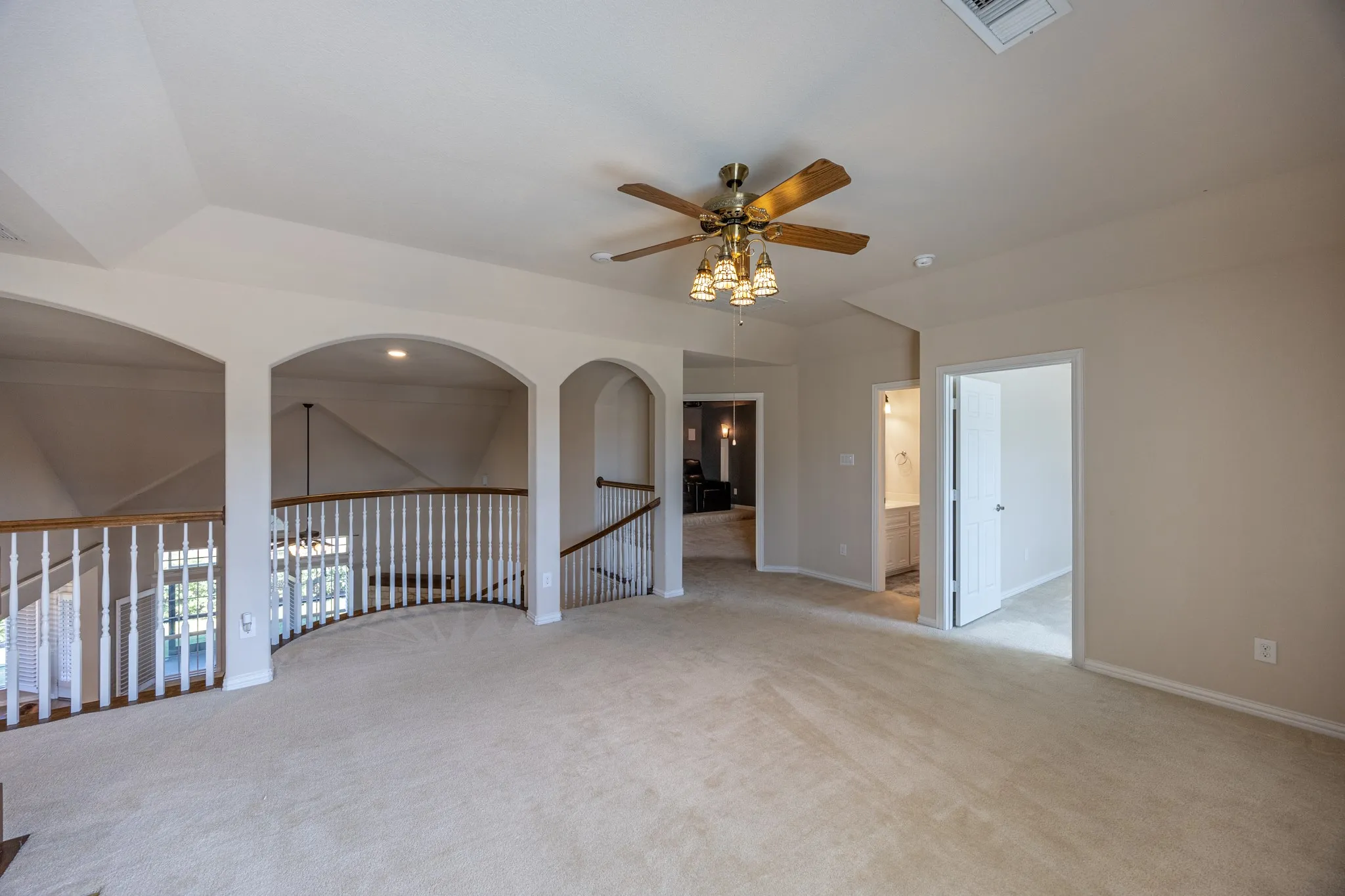 Spacious loft area featuring neutral carpet flooring, arched wall openings, a brass-finish ceiling fan with lighting, and white baluster railings with wood-finish handrails