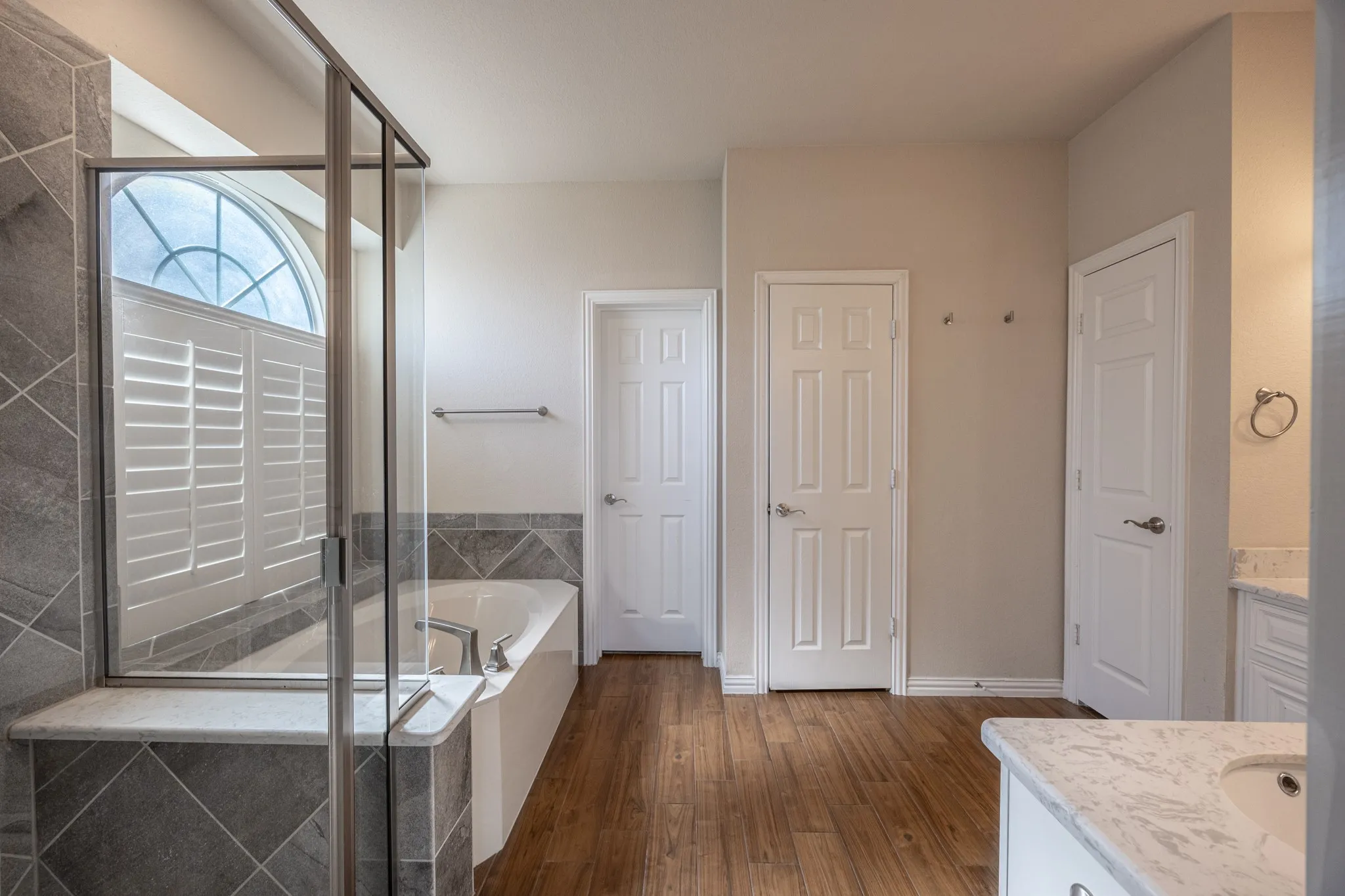 Bathroom featuring a deep soaking tub with gray tile surround, glass-enclosed shower, wood-finish flooring, plantation shutters on an arched window, and a vanity with a white quartz countertop